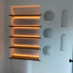 Stepan Malitskyi Standem Bud - Biała ściana z podświetlanymi półkami z drewna i wnękami w kształcie kół i prostokątów. Widoczny kabel elektryczny i zabezpieczona podłoga.