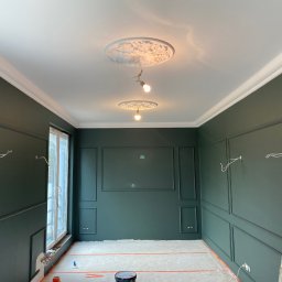 Stepan Malitskyi Standem Bud - Eleganckie wnętrze w trakcie malowania na ciemnozielono, z dekoracyjnymi listwami ściennymi i rozetami sufitowymi. Widoczna drabina i zabezpieczona podłoga.