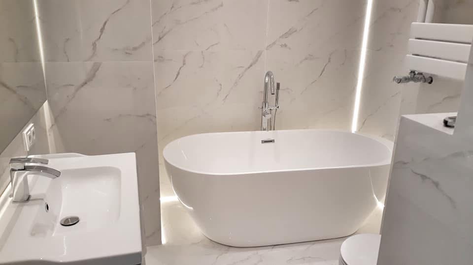 Biała, wolnostojąca wanna w nowoczesnej łazience z marmurowymi płytkami i subtelnym oświetleniem LED. Umywalka i grzejnik drabinkowy dopełniają minimalistyczny wystrój.