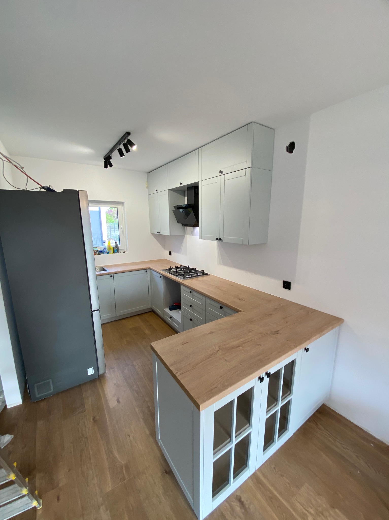 Jasna kuchnia po remoncie: białe ściany, drewniana podłoga, szare meble z drewnianym blatem. Nowoczesne oświetlenie punktowe. Widoczna płyta gazowa i szary lodówka.