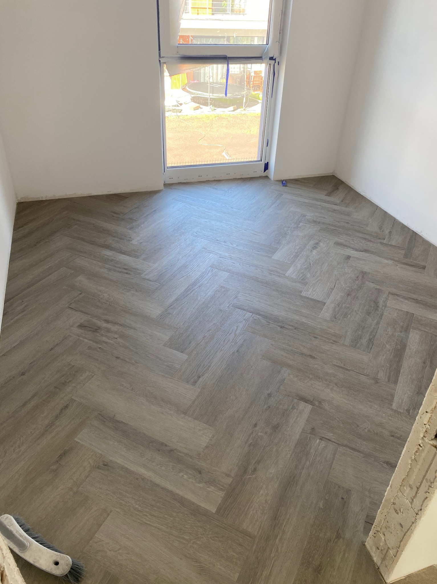 Ułożona podłoga z paneli w jodełkę w jasnym pokoju z oknem. Widoczne niedoskonałości przy ścianie i narzędzie pracy w rogu.