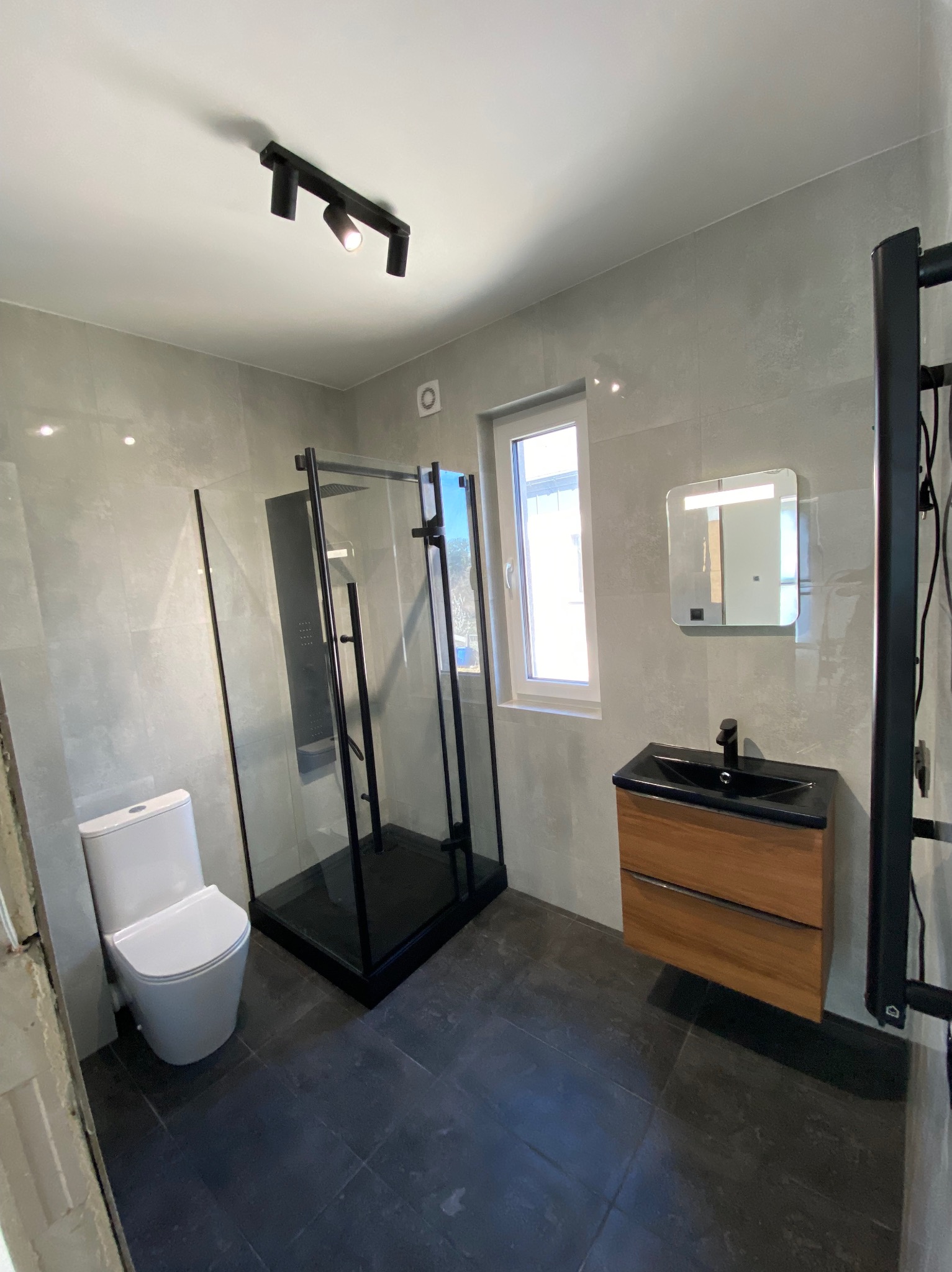 Wykończona łazienka z czarną kabiną prysznicową, umywalką z drewnianą szafką, toaletą i grzejnikiem drabinkowym. Szare płytki na ścianach i podłodze. Nowoczesne oświetlenie.