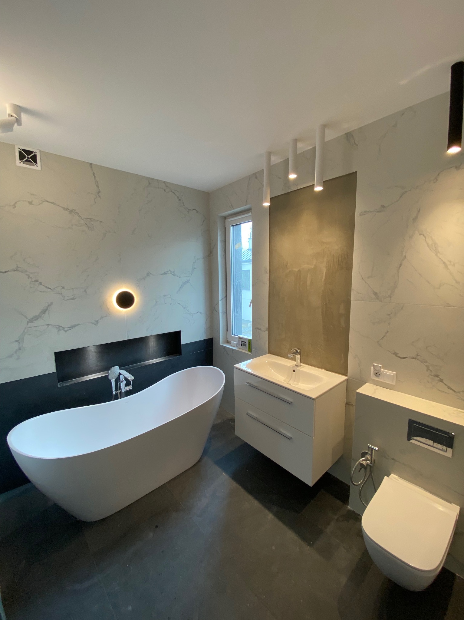 Nowoczesna łazienka z wolnostojącą wanną, umywalką z szafką i podwieszaną toaletą. Marmurowe płytki na ścianach, czarna podłoga i oświetlenie punktowe.