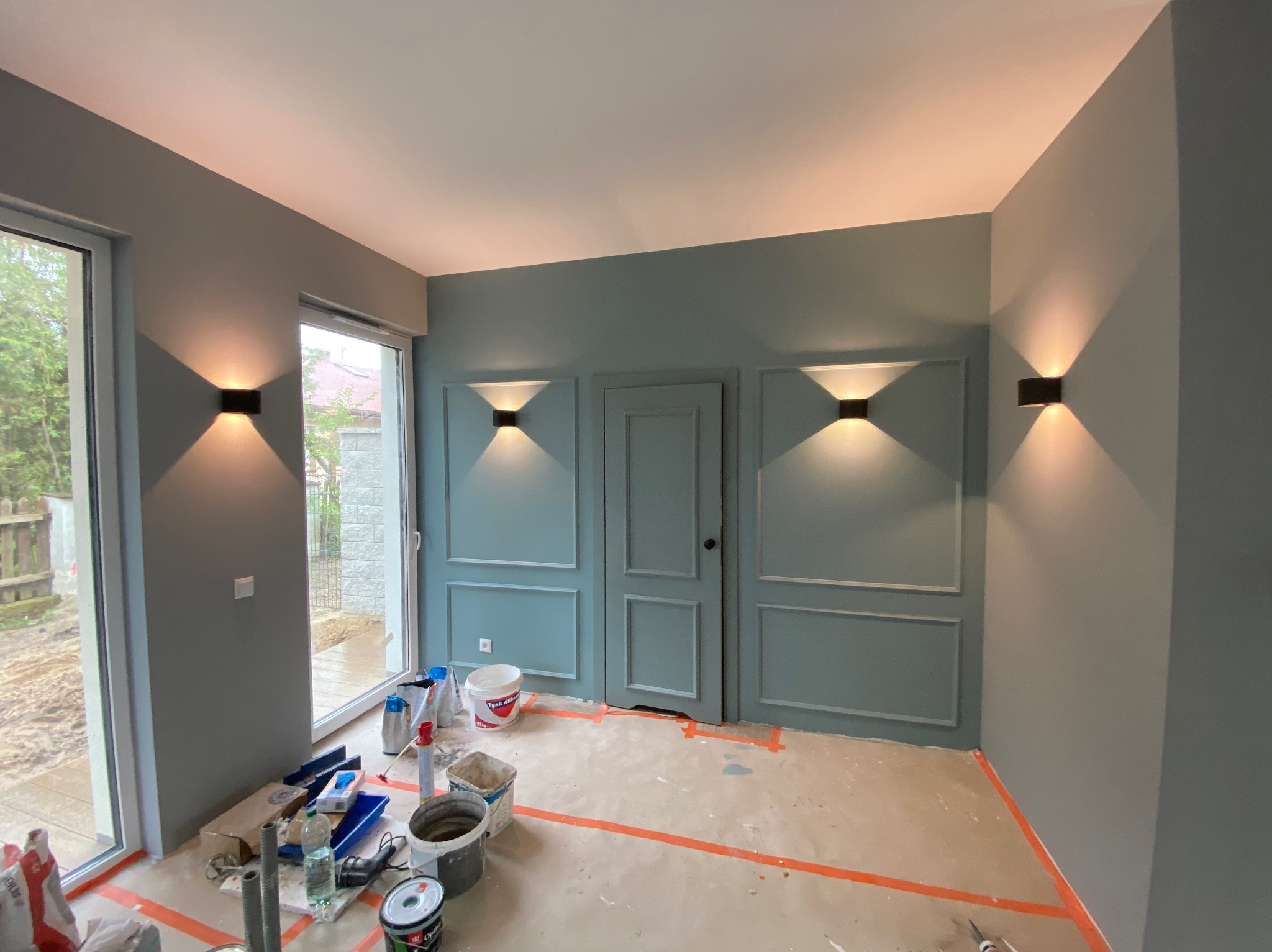 Pokój po malowaniu w odcieniach szarości i błękitu z dekoracyjnymi listwami i nowoczesnym oświetleniem ściennym. Widoczne narzędzia malarskie.