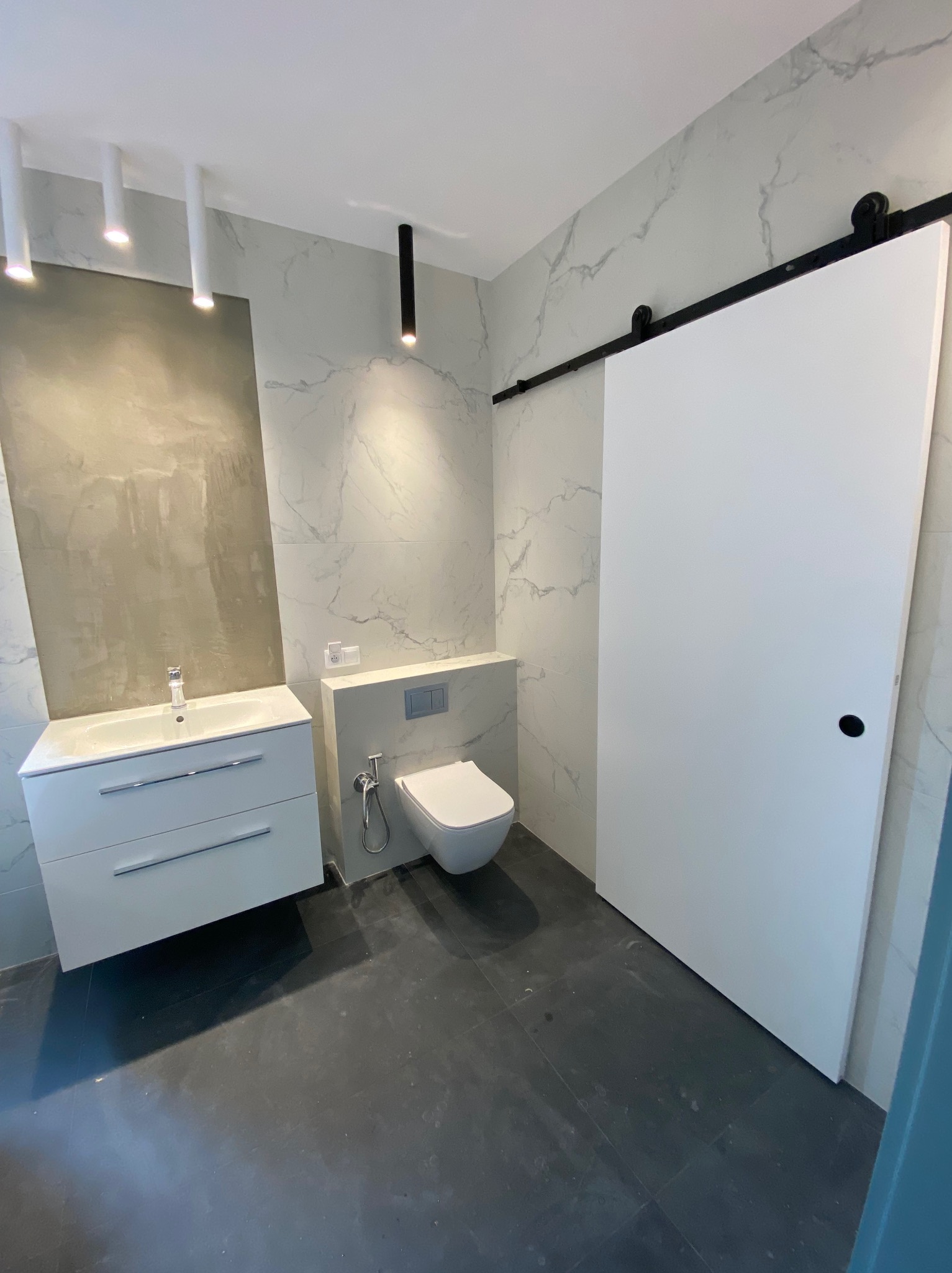 Nowoczesna łazienka z marmurowymi płytkami, wiszącą toaletą, białą szafką z umywalką i przesuwnymi drzwiami na szynie w kolorze czarnym.