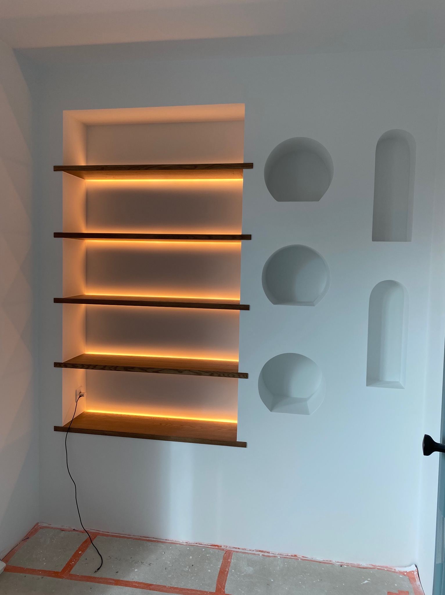 Biała ściana z podświetlanymi półkami z drewna i wnękami w kształcie kół i prostokątów. Widoczny kabel elektryczny i zabezpieczona podłoga.