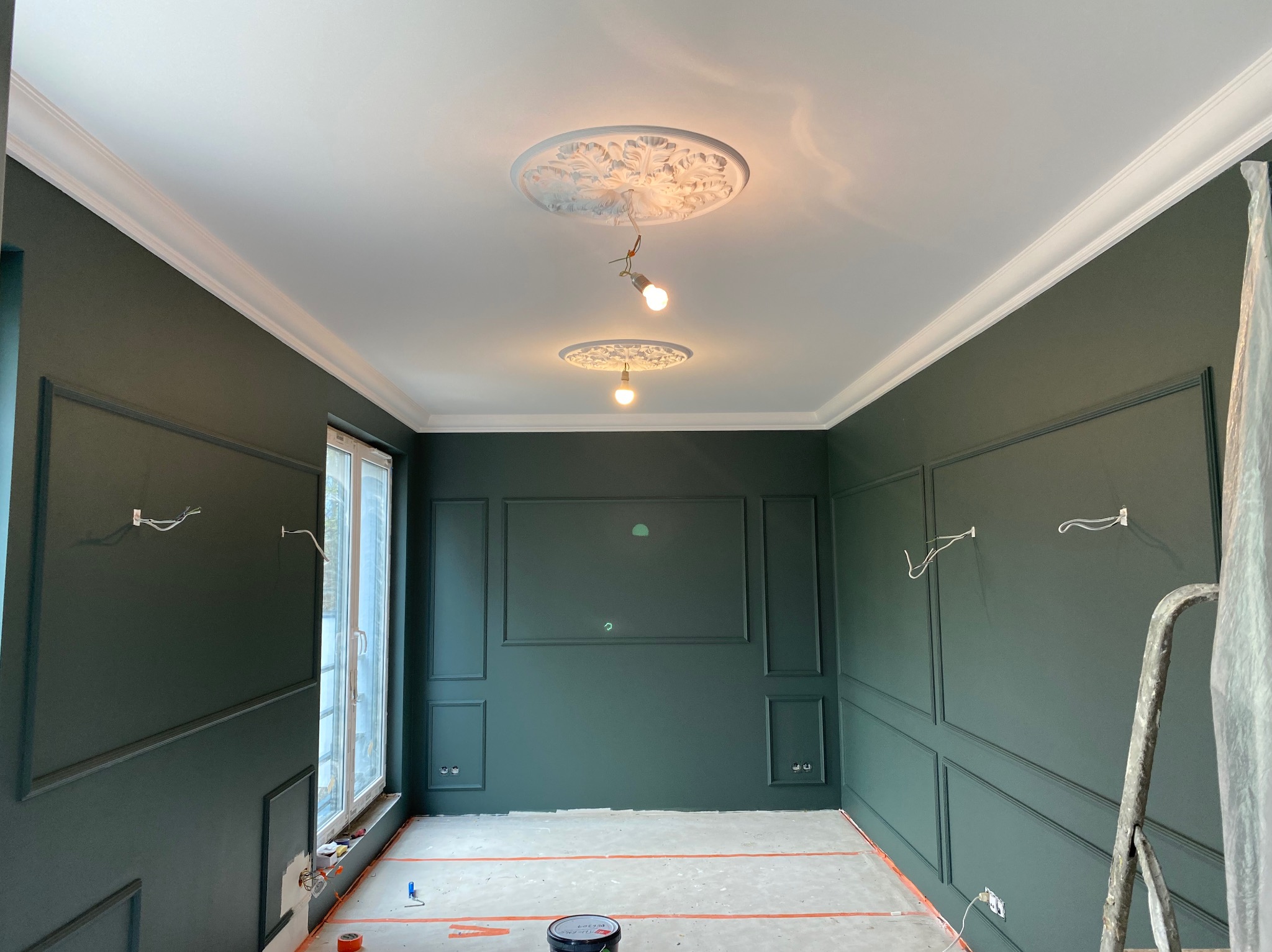 Eleganckie wnętrze w trakcie malowania na ciemnozielono, z dekoracyjnymi listwami ściennymi i rozetami sufitowymi. Widoczna drabina i zabezpieczona podłoga.