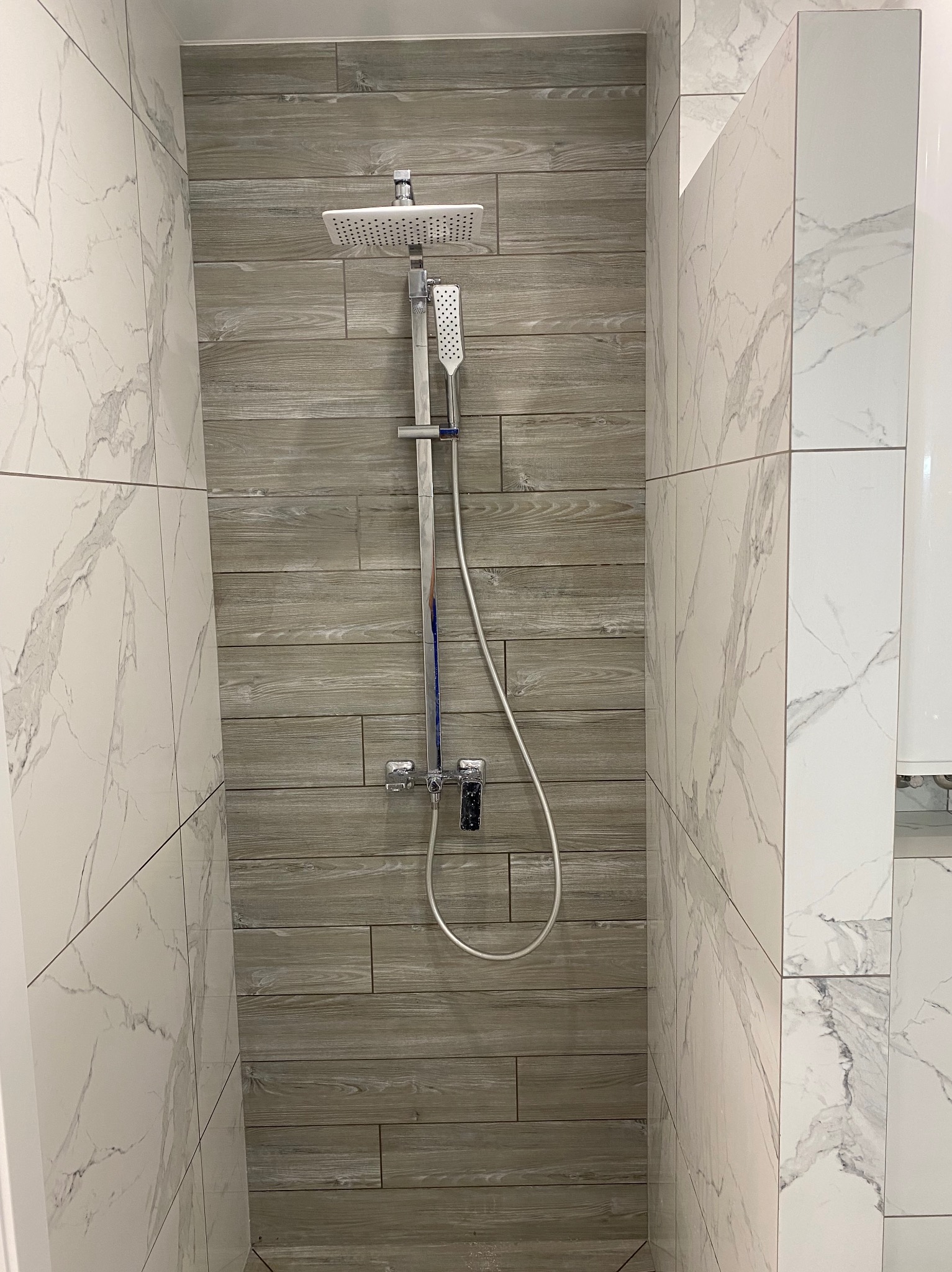 Kabina prysznicowa z szarą armaturą i płytkami imitującymi drewno na ścianie, obok białe płytki z wzorem marmuru. Nowoczesny design.