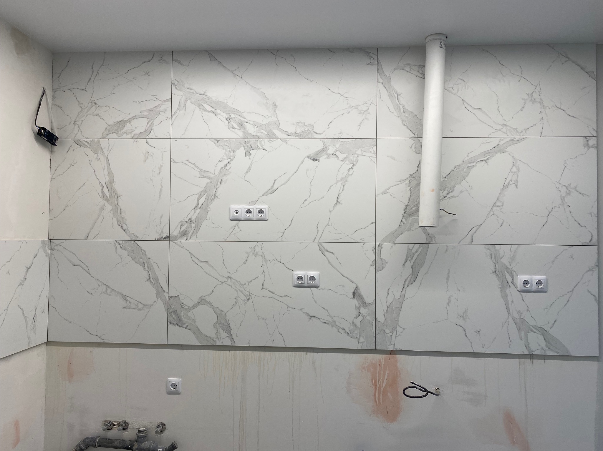 Wykończona ściana w kuchni z białych płytek imitujących marmur, widoczne gniazdka elektryczne i fragment instalacji wentylacyjnej. Ściana częściowo przygotowana do dalszych prac.