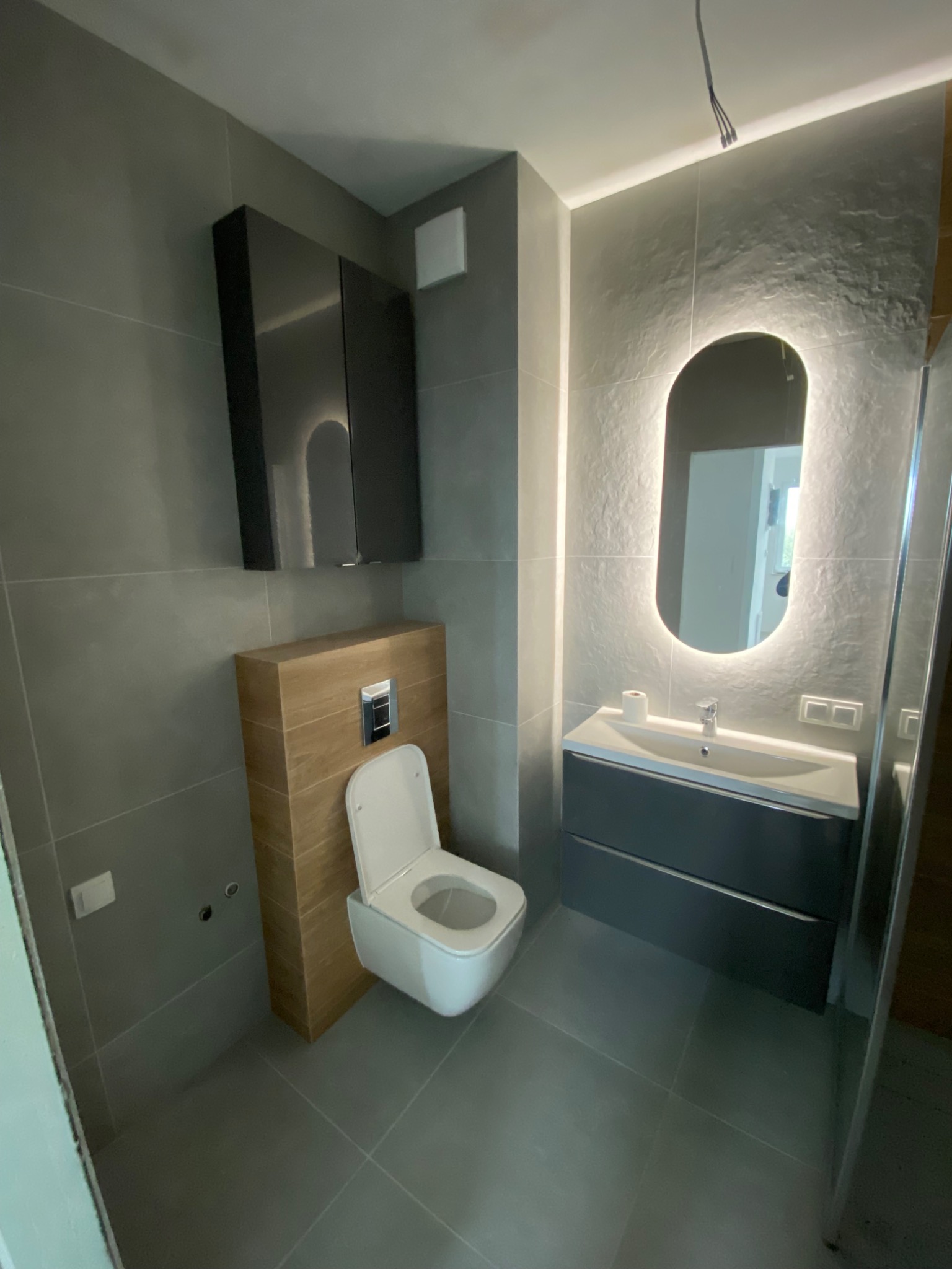 Nowoczesna łazienka z szarą glazurą, podświetlanym lustrem i podwieszaną toaletą z drewnianą obudową. Minimalistyczny design, eleganckie wykończenie.