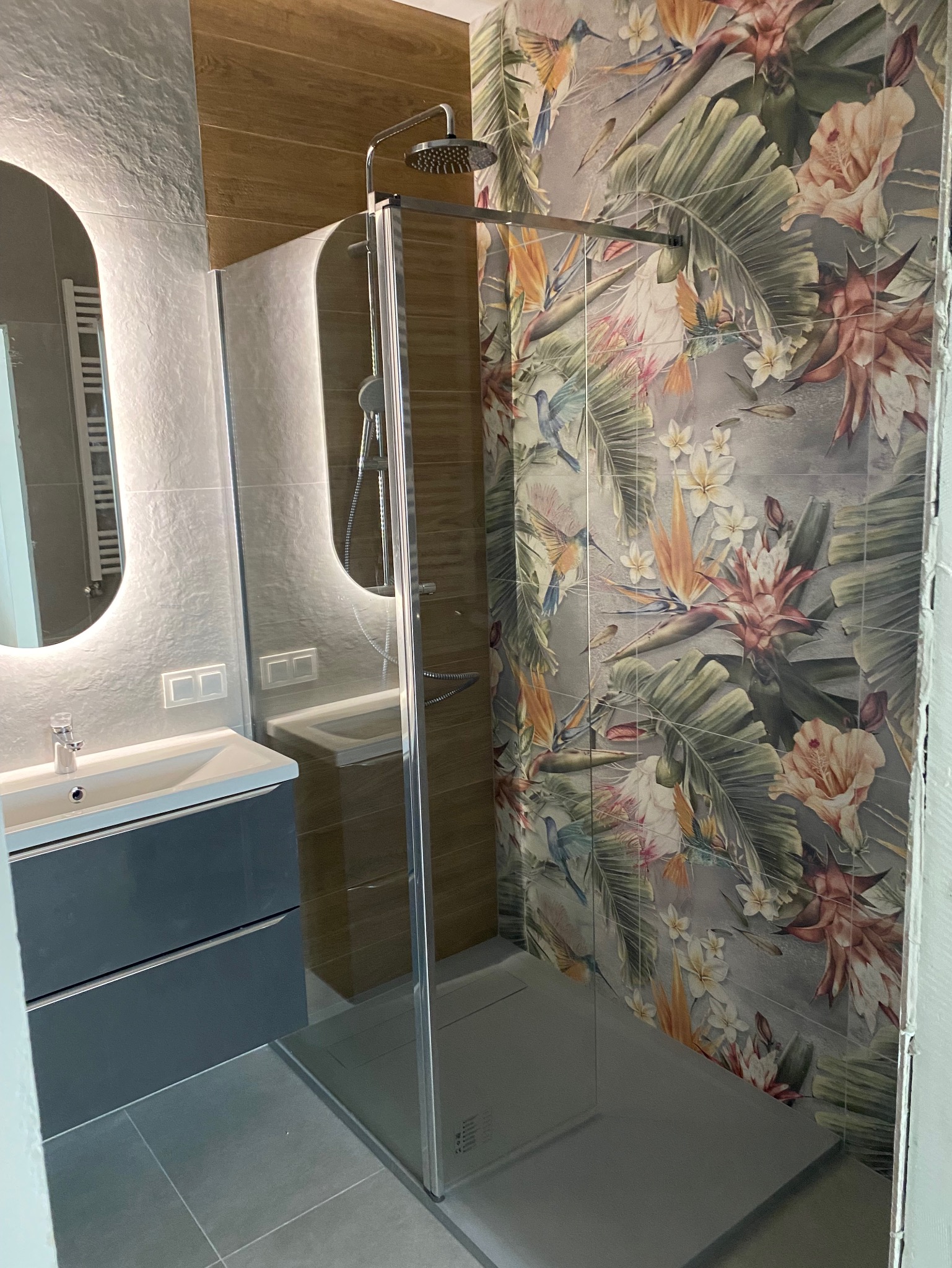 Wykończona łazienka z kabiną prysznicową typu walk-in, umywalką z szafką w odcieniach szarości i płytkami z motywem tropikalnym na ścianie.