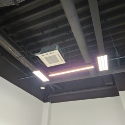 Nsinstal klimatyzacja wentylacja rekuperacja - Sufit z zamontowaną kasetonową klimatyzacją, widoczne rury wentylacyjne i oświetlenie liniowe. Czarny sufit kontrastuje z białymi ścianami.