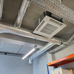 Nsinstal klimatyzacja wentylacja rekuperacja - Zamontowana klimatyzacja kasetonowa Fujitsu na suficie, widoczne srebrne rury wentylacyjne i metalowe korytka kablowe. Ujęcie z dołu, fragment regału w rogu.