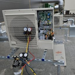 Nsinstal klimatyzacja wentylacja rekuperacja - Otwarta jednostka zewnętrzna klimatyzacji Fujitsu na dachu budynku, podłączona do manometrów i butli z gazem, w tle widoczne inne jednostki i elementy instalacji.