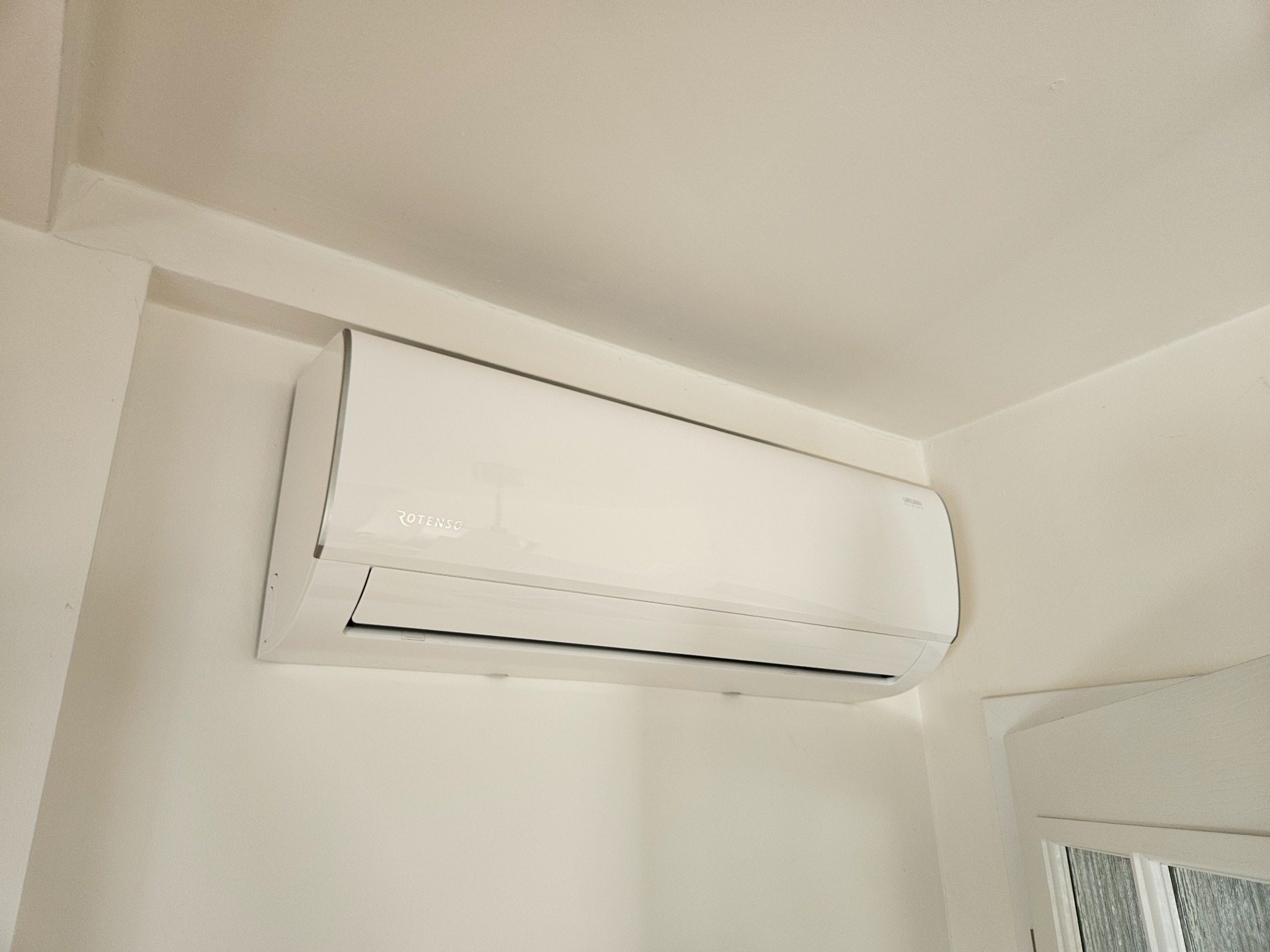Biała klimatyzacja Rotenso zamontowana na ścianie w jasnym pomieszczeniu, widoczna z dołu pod kątem. Minimalistyczny design, czyste linie, nowoczesny wygląd.