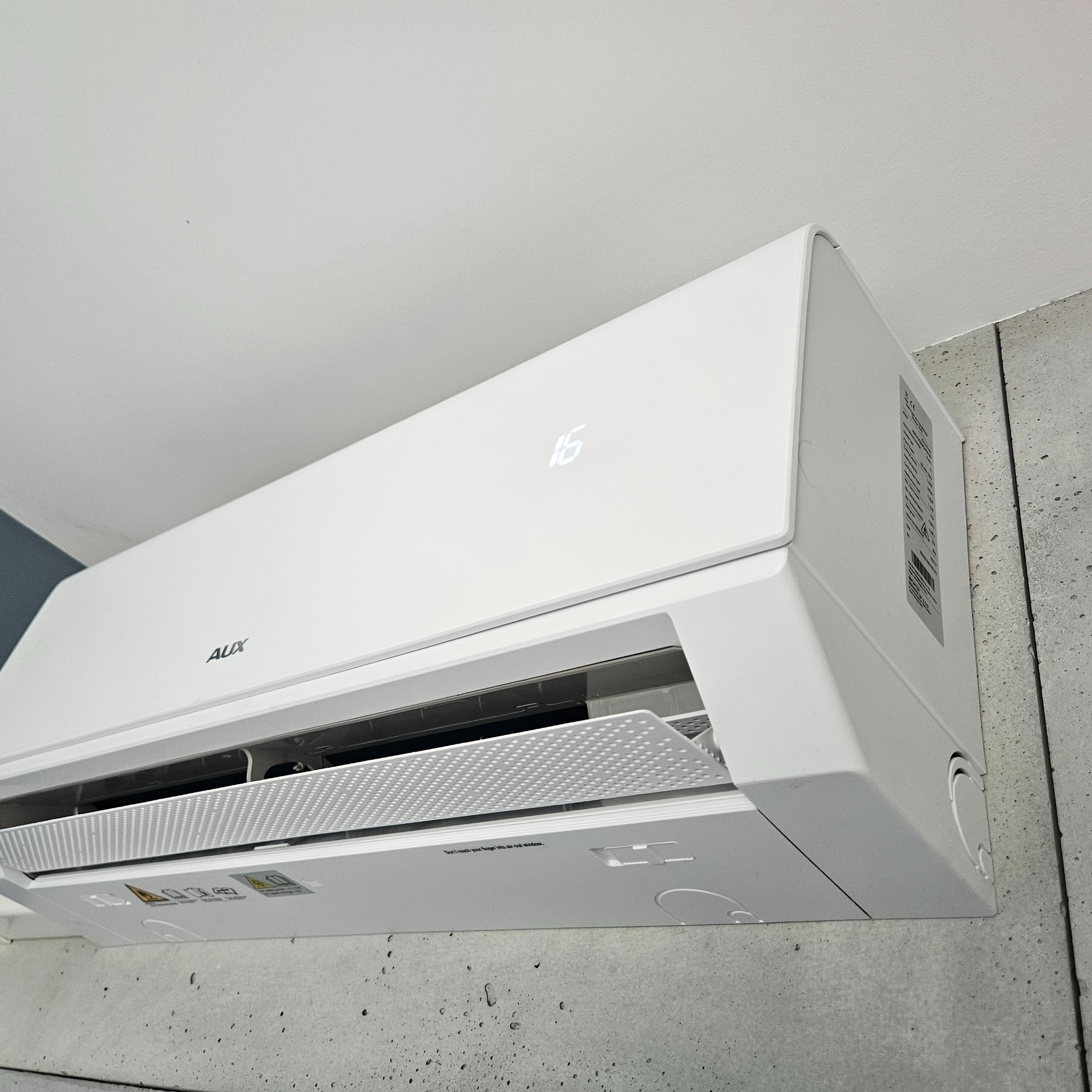 Biała klimatyzacja AUX na ścianie z betonu architektonicznego, widoczny wyświetlacz z temperaturą 16 stopni. Ujęcie z dołu, lekko z prawej strony.