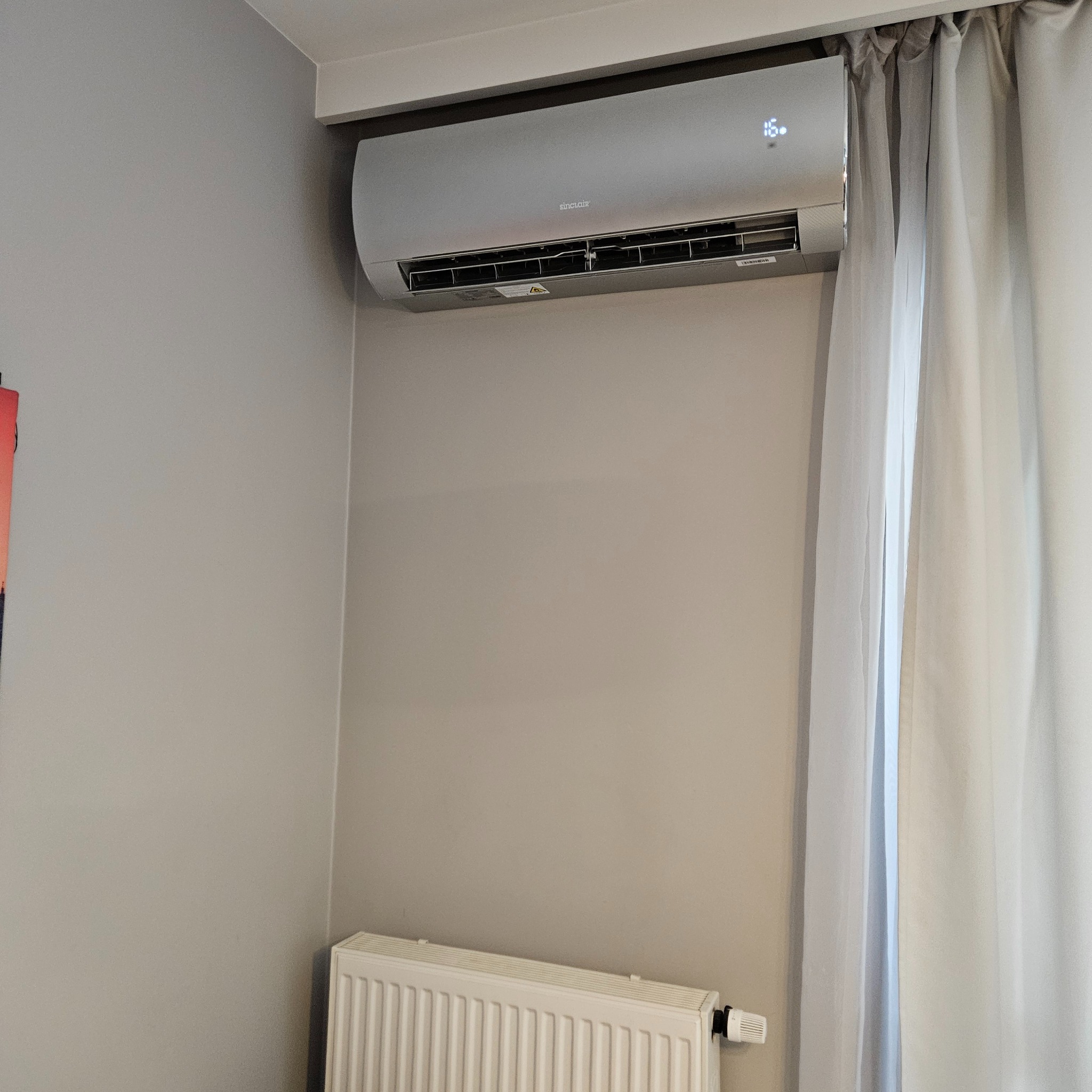 Nowoczesny, srebrny klimatyzator Sinclair zamontowany na szarej ścianie w pokoju. Widoczny biały grzejnik pod klimatyzatorem i zasłona w tle. Wyświetlacz pokazuje 16 stopni.