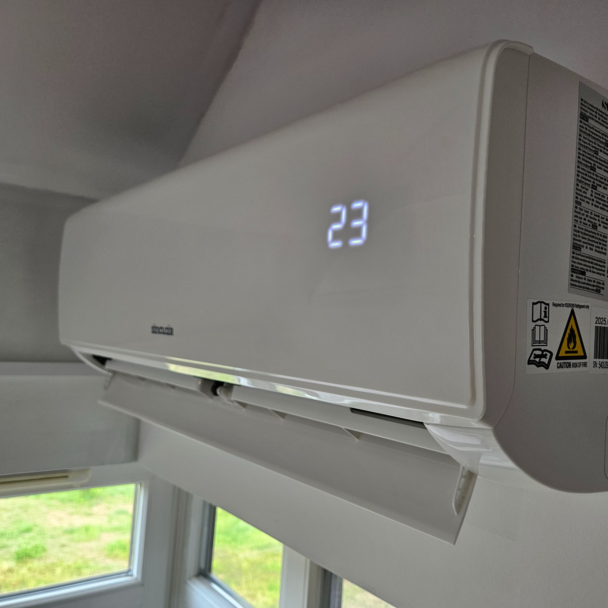 Biała klimatyzacja Sinclair z wyświetlaczem temperatury 23 stopnie, zamontowana w pomieszczeniu z oknem, widok z dołu, etykieta ostrzegawcza po prawej stronie.