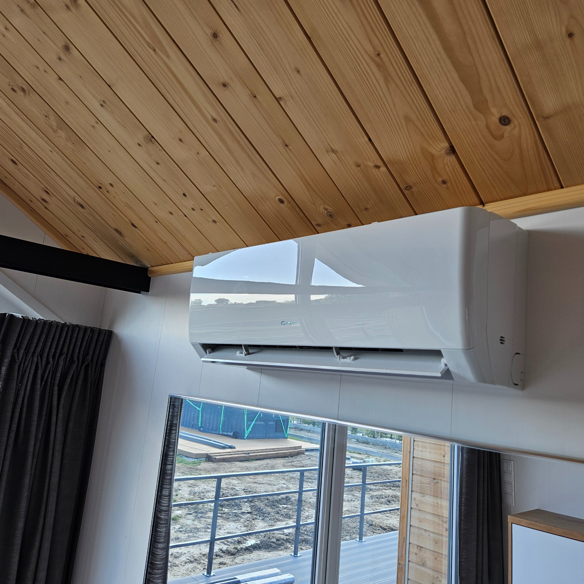 Biały klimatyzator ścienny marki Gree zamontowany w pomieszczeniu z drewnianym sufitem i widokiem na zewnątrz przez okno. Widoczne odbicie w obudowie.