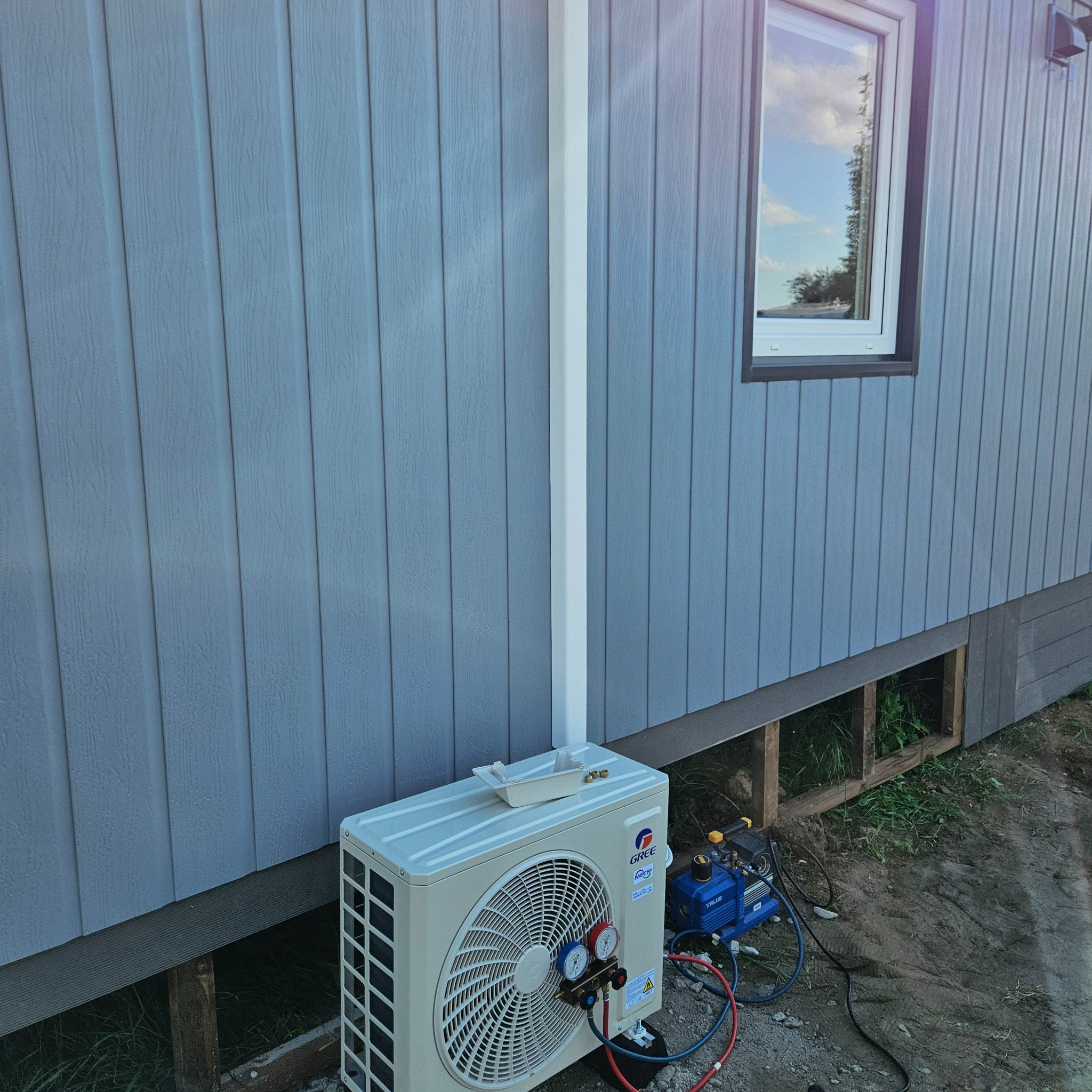 Biała jednostka zewnętrzna klimatyzacji Gree zamontowana przy błękitnej elewacji domu z widocznym oknem i narzędziami serwisowymi obok.