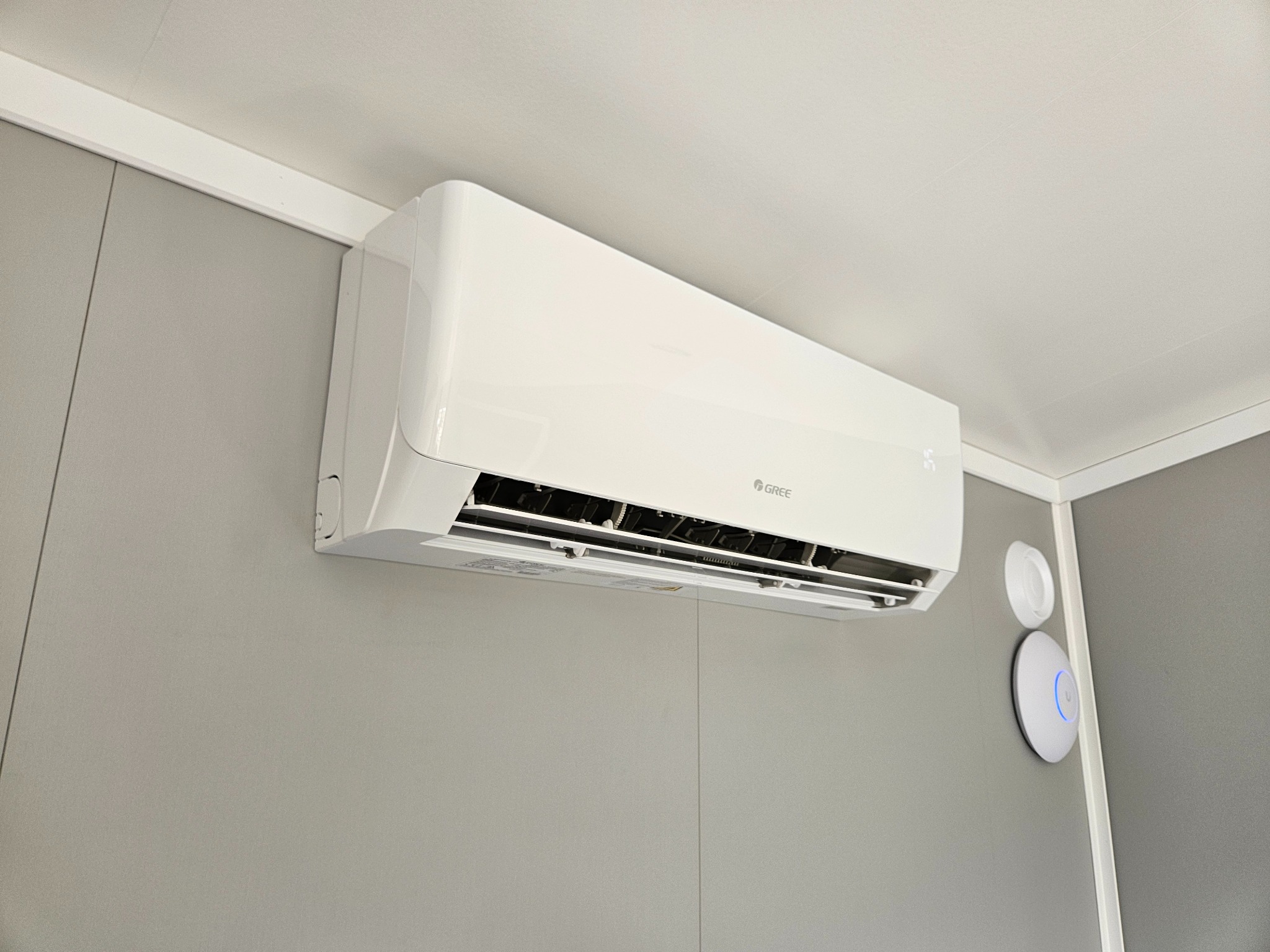 Biała klimatyzacja Gree na szarej ścianie, widoczny czujnik temperatury. Minimalistyczny design, nowoczesne wnętrze, oświetlenie dzienne, kąt ujęcia od dołu.