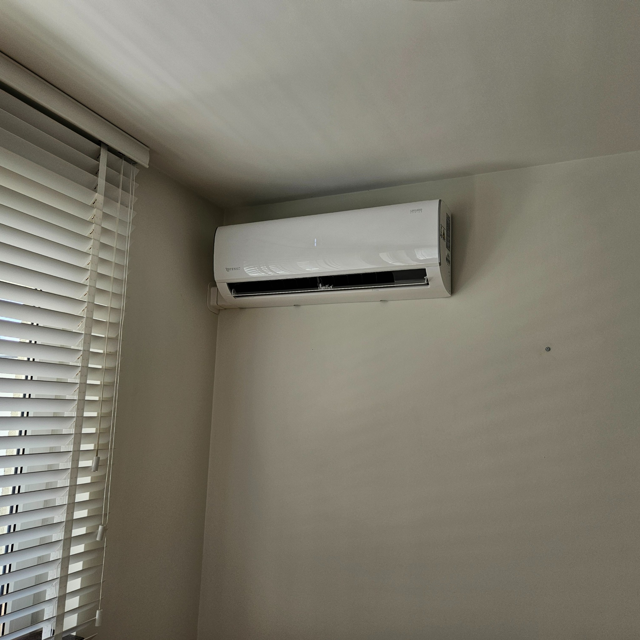Biała klimatyzacja Rotenso zamontowana na ścianie w pokoju z żaluzjami. Widok z dołu, oświetlenie dzienne.