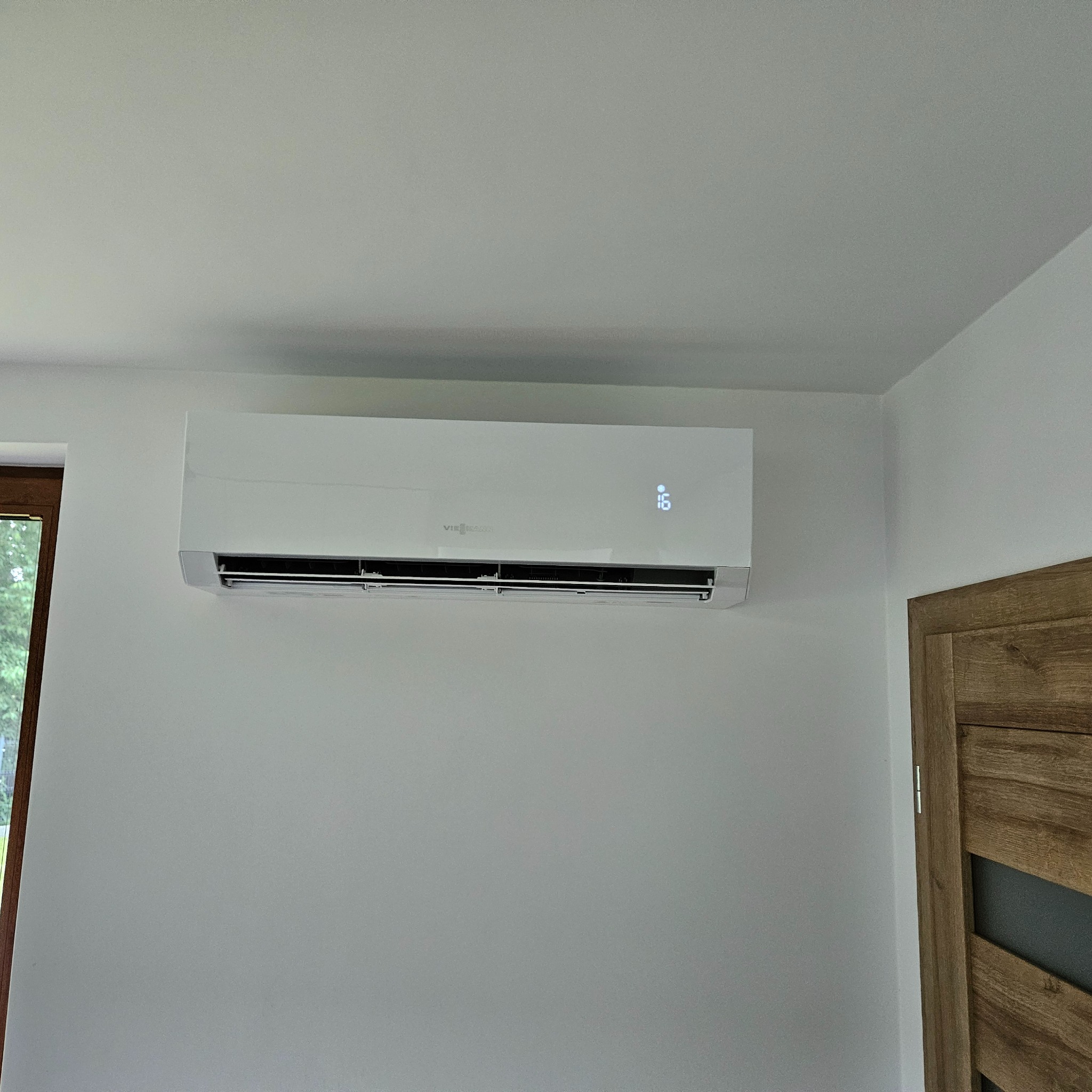 Biała klimatyzacja Viessmann na ścianie w jasnym wnętrzu, widoczny fragment okna i drzwi. Minimalistyczny design, nowoczesne rozwiązanie chłodnicze.