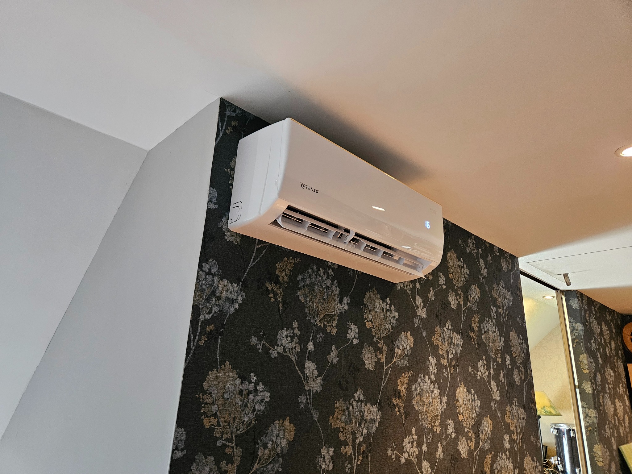 Biały klimatyzator Rotenso zamontowany na ścianie z ciemną tapetą w kwiaty. Widoczny wyświetlacz temperatury. Ujęcie z dołu.