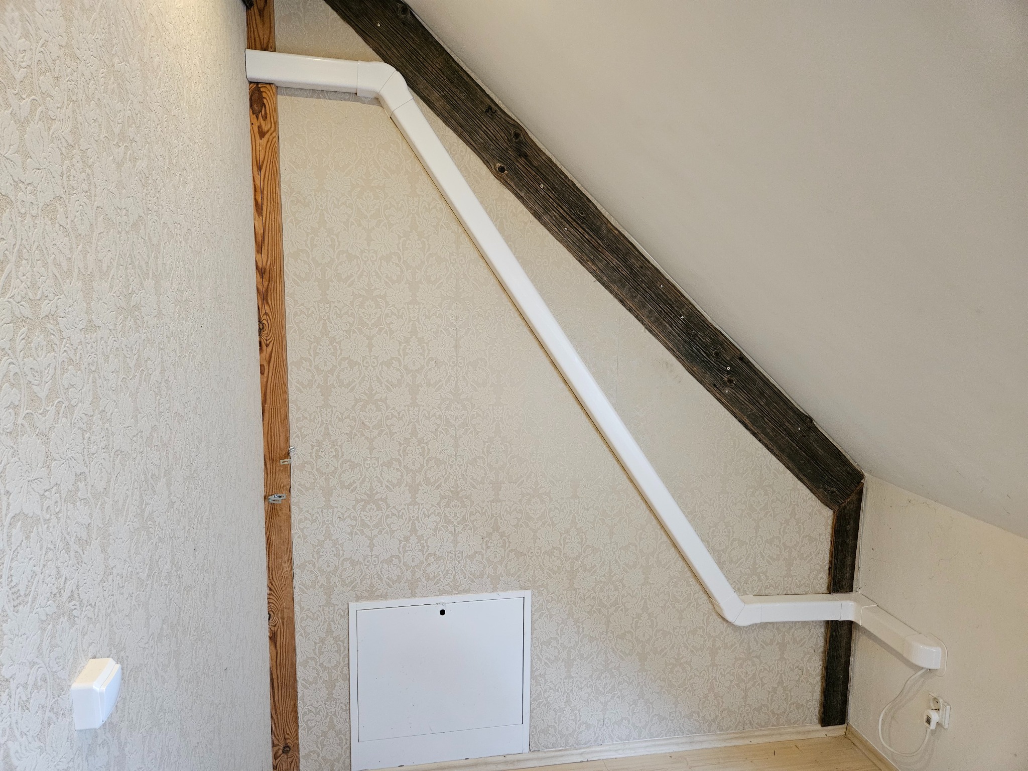 Białe rury wentylacyjne poprowadzone po skosie poddasza, widoczne drewniane elementy konstrukcyjne i wzorzysta tapeta. Instalacja w trakcie montażu.