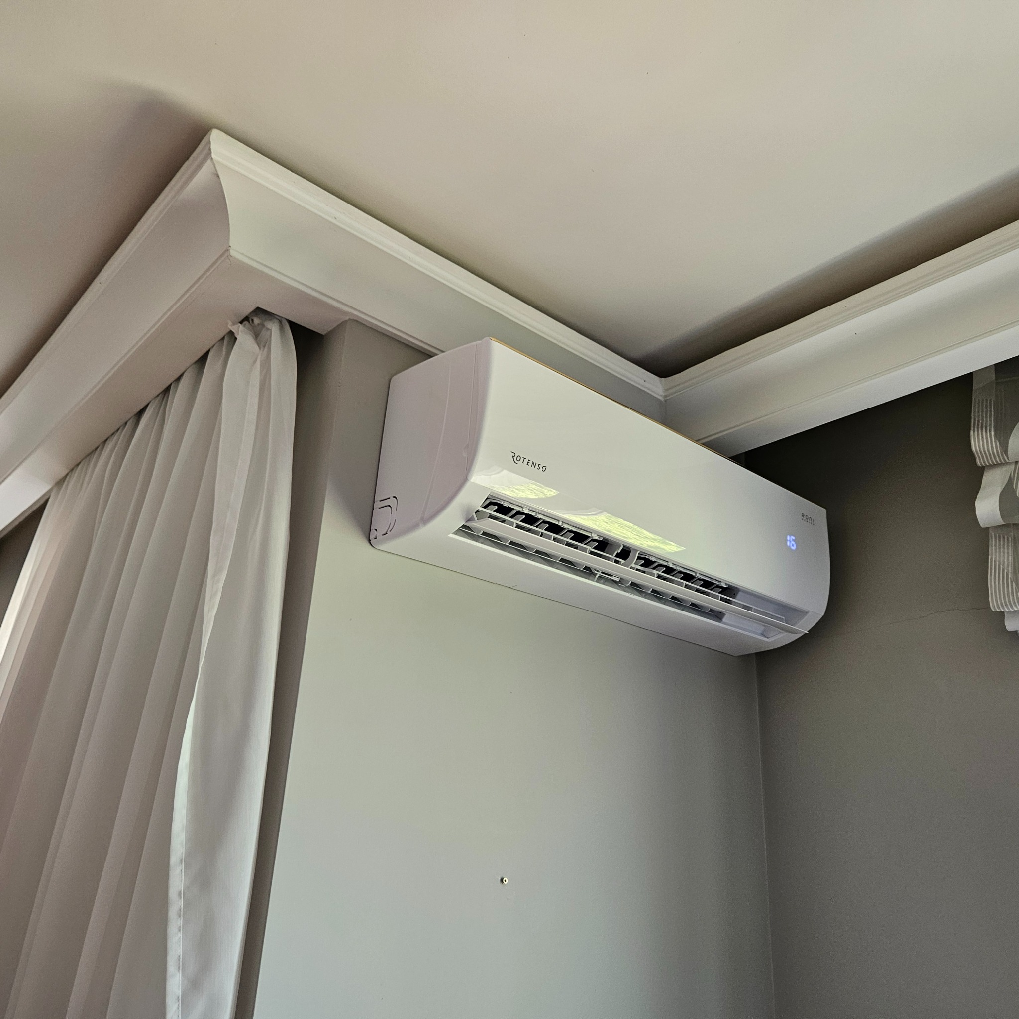 Biały klimatyzator Rotenso zamontowany na ścianie w pokoju z zasłonami i ozdobną listwą sufitową. Widoczny wyświetlacz z temperaturą 16 stopni.