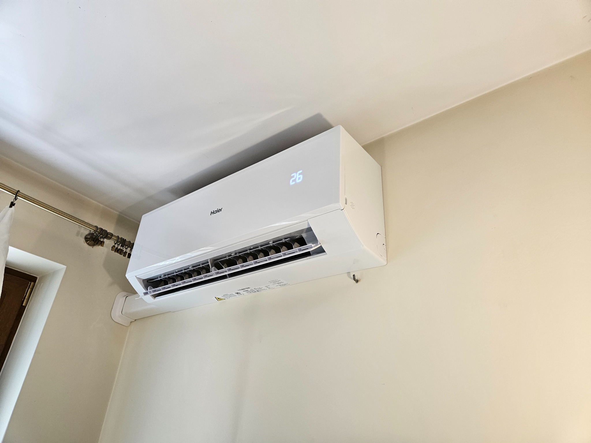 Biała klimatyzacja Haier z wyświetlaczem 26 stopni Celsjusza, zamontowana na beżowej ścianie w jasnym wnętrzu, widoczna z dołu pod kątem.