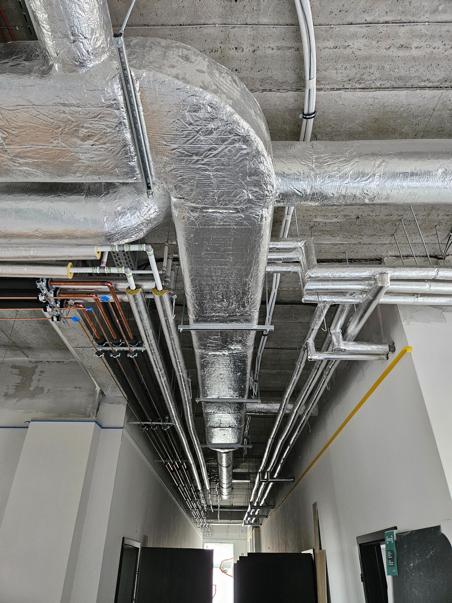 Srebrne, zaizolowane kanały wentylacyjne i rury pod sufitem w korytarzu budynku komercyjnego. Widoczne elementy instalacji miedzianej i stalowej.