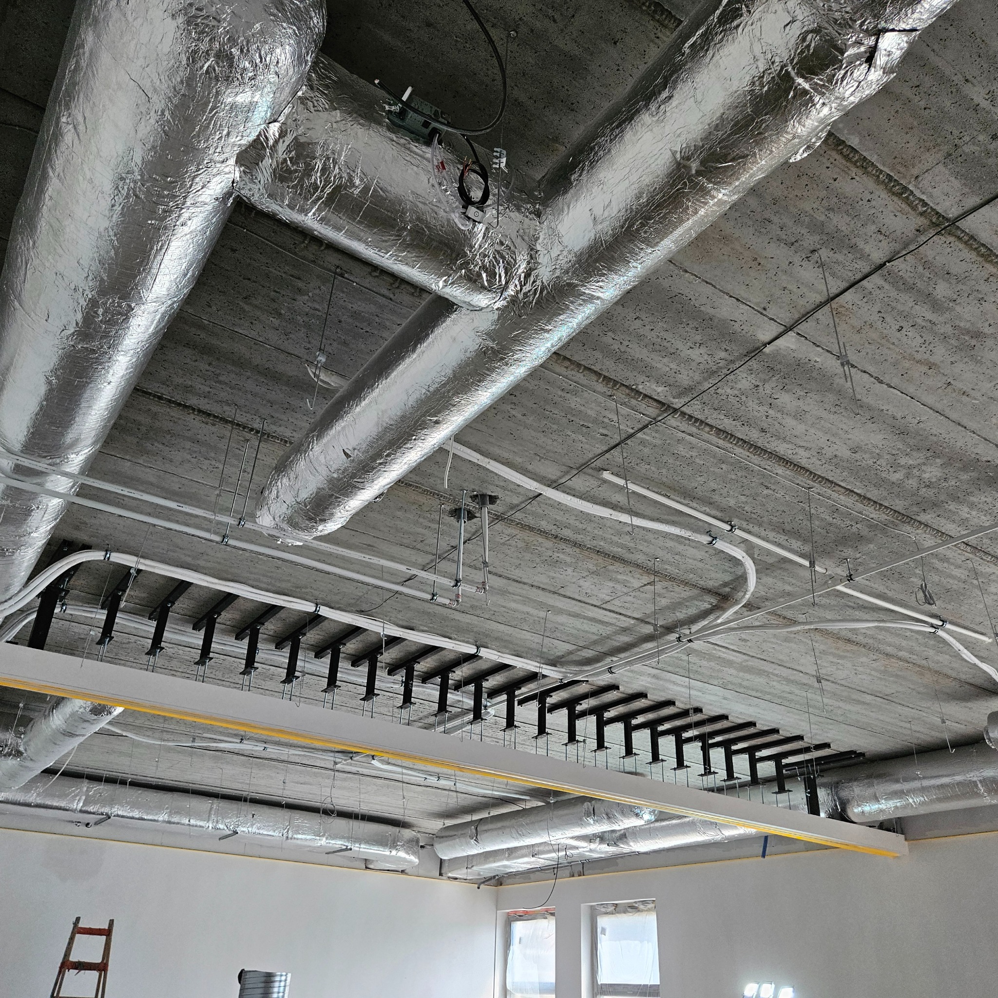 Srebrne, zaizolowane rury wentylacyjne pod betonowym sufitem, widoczne elementy instalacji i okna w tle. Ujęcie z dołu, szeroki kadr.