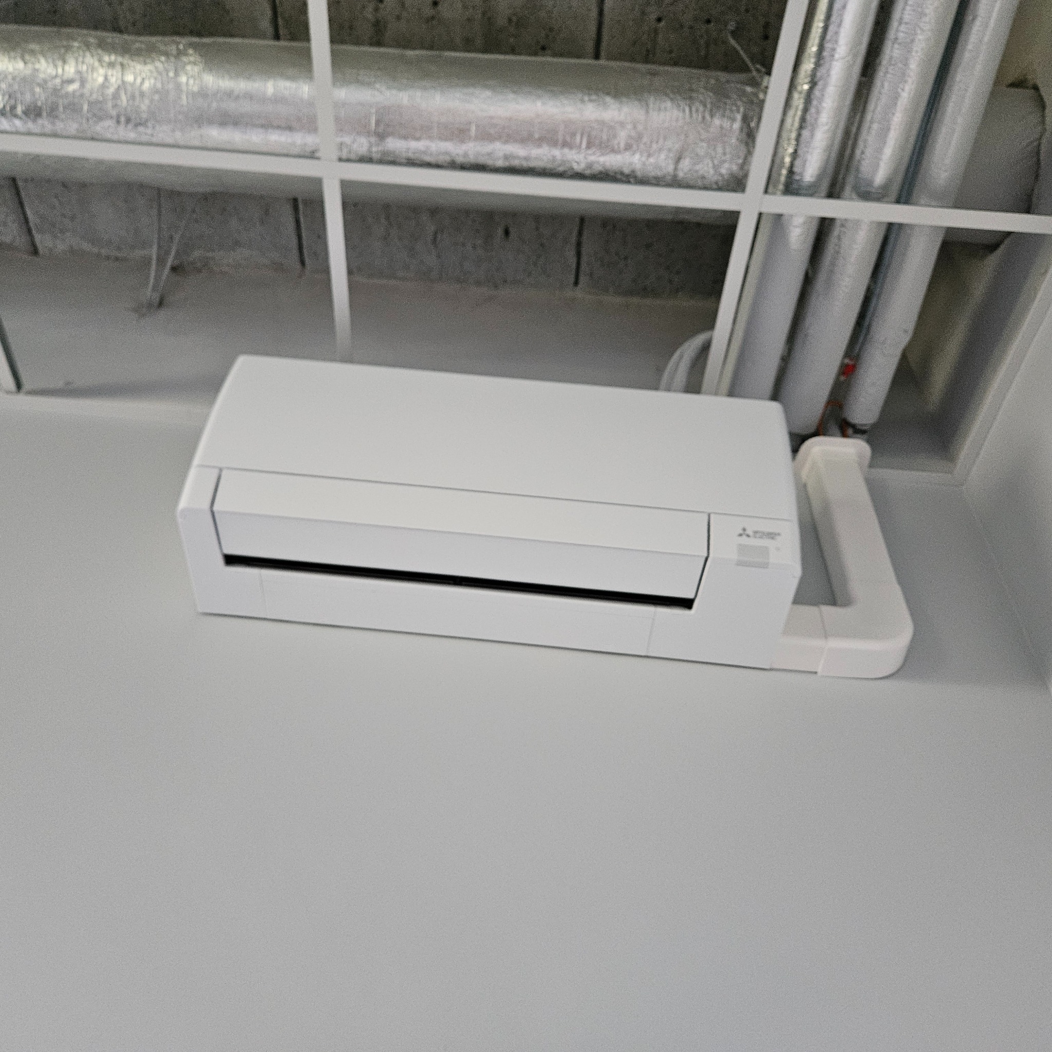 Biała jednostka wewnętrzna klimatyzacji Mitsubishi Electric zamontowana pod sufitem z widocznymi rurami instalacyjnymi i elementami konstrukcyjnymi.