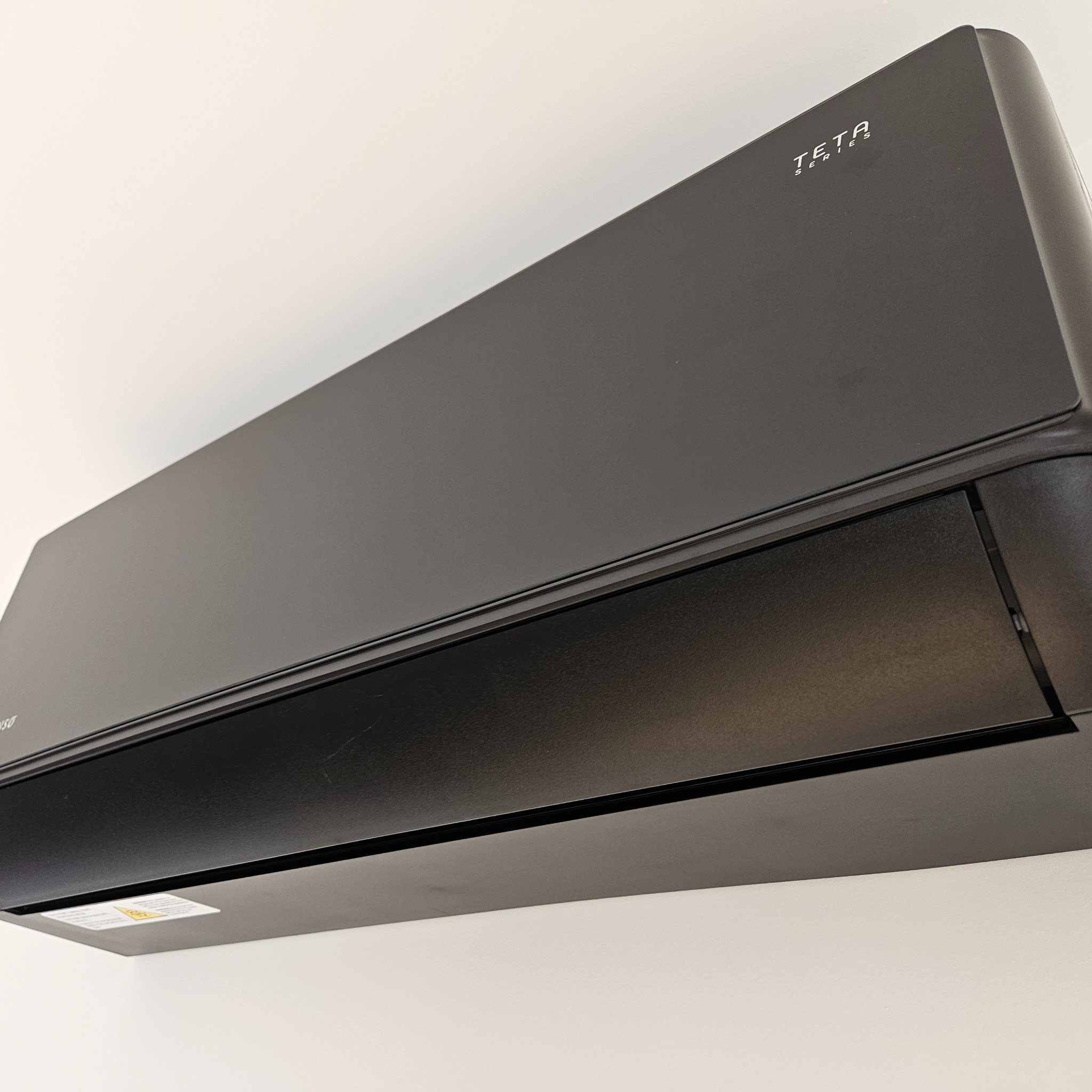 Czarny klimatyzator TETA Series zamontowany na białej ścianie, widok z dołu, ukazujący nowoczesny design i minimalistyczny styl urządzenia.