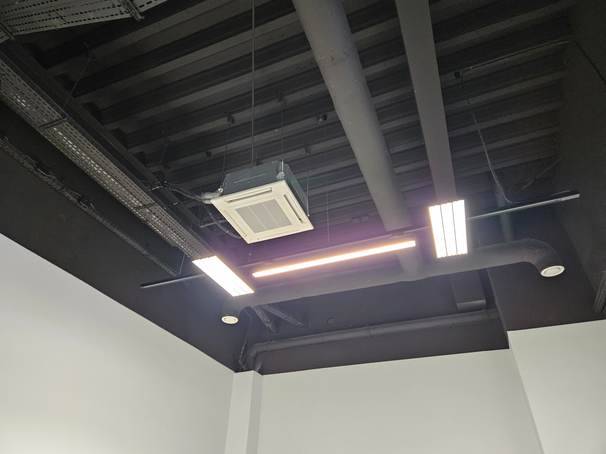 Sufit z zamontowaną kasetonową klimatyzacją, widoczne rury wentylacyjne i oświetlenie liniowe. Czarny sufit kontrastuje z białymi ścianami.