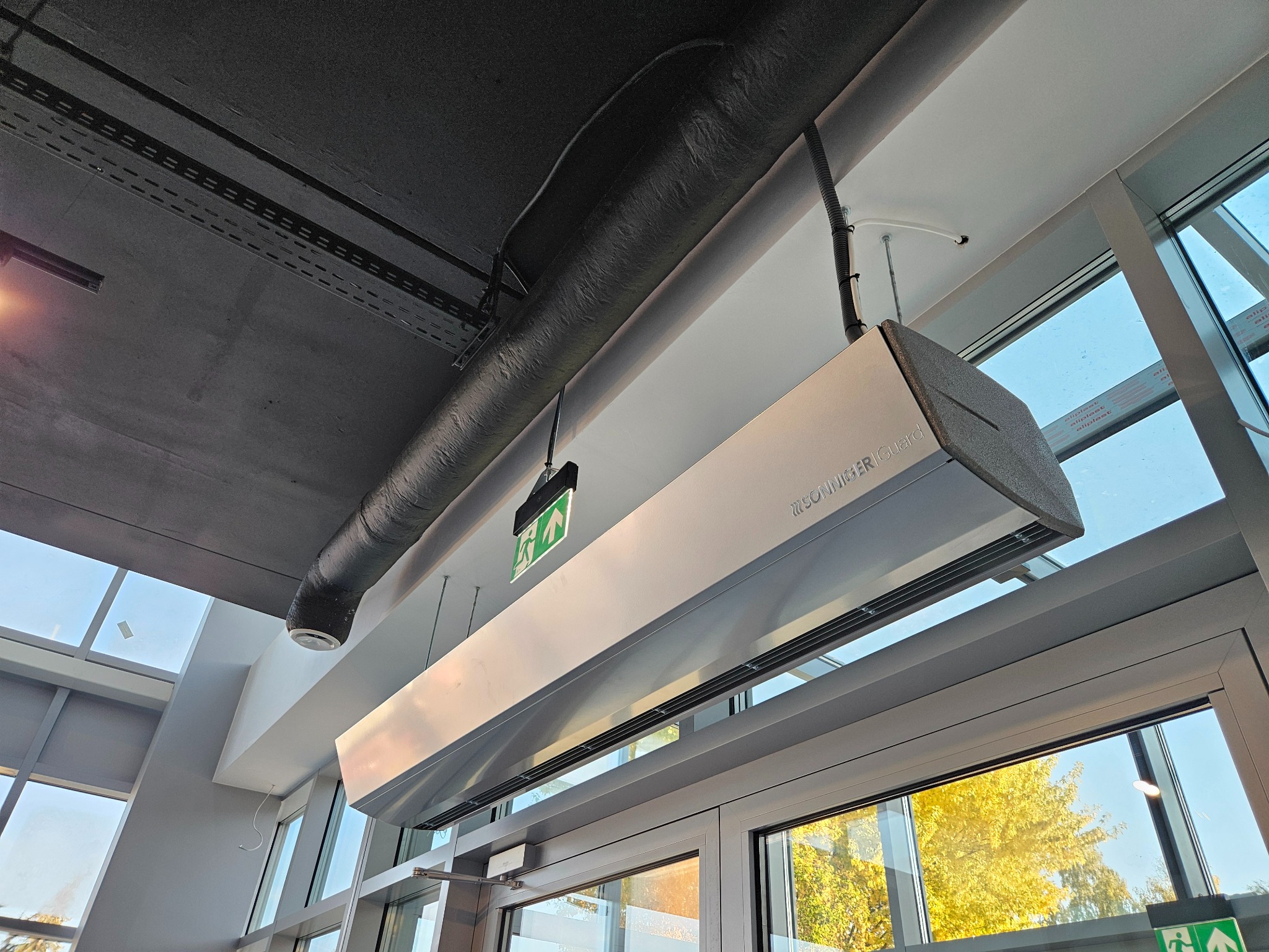 Sufitowa kurtyna powietrzna Sonniger Guard nad wejściem, widoczna instalacja, rura wentylacyjna i znak ewakuacyjny. Jasne wnętrze z widokiem na zewnątrz.