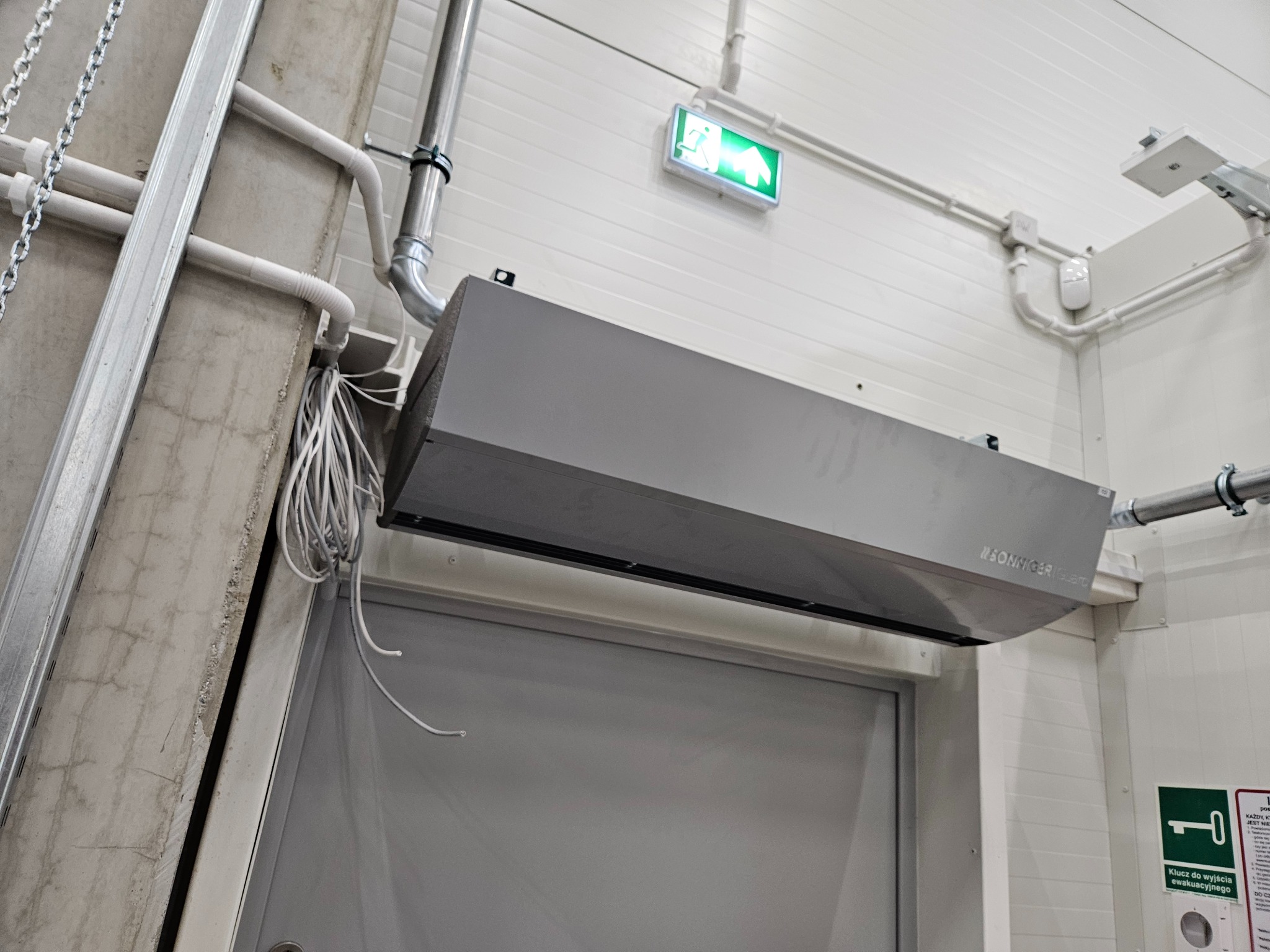 Szara kurtyna powietrzna marki Sonniger Guard zamontowana nad drzwiami w pomieszczeniu o białych ścianach, widoczna instalacja i znak ewakuacyjny.