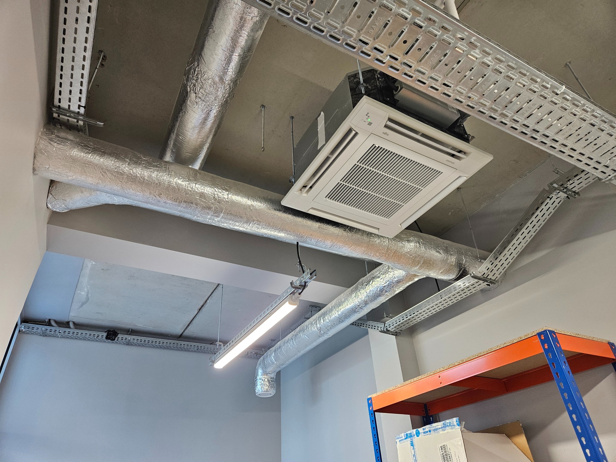 Zamontowana klimatyzacja kasetonowa Fujitsu na suficie, widoczne srebrne rury wentylacyjne i metalowe korytka kablowe. Ujęcie z dołu, fragment regału w rogu.