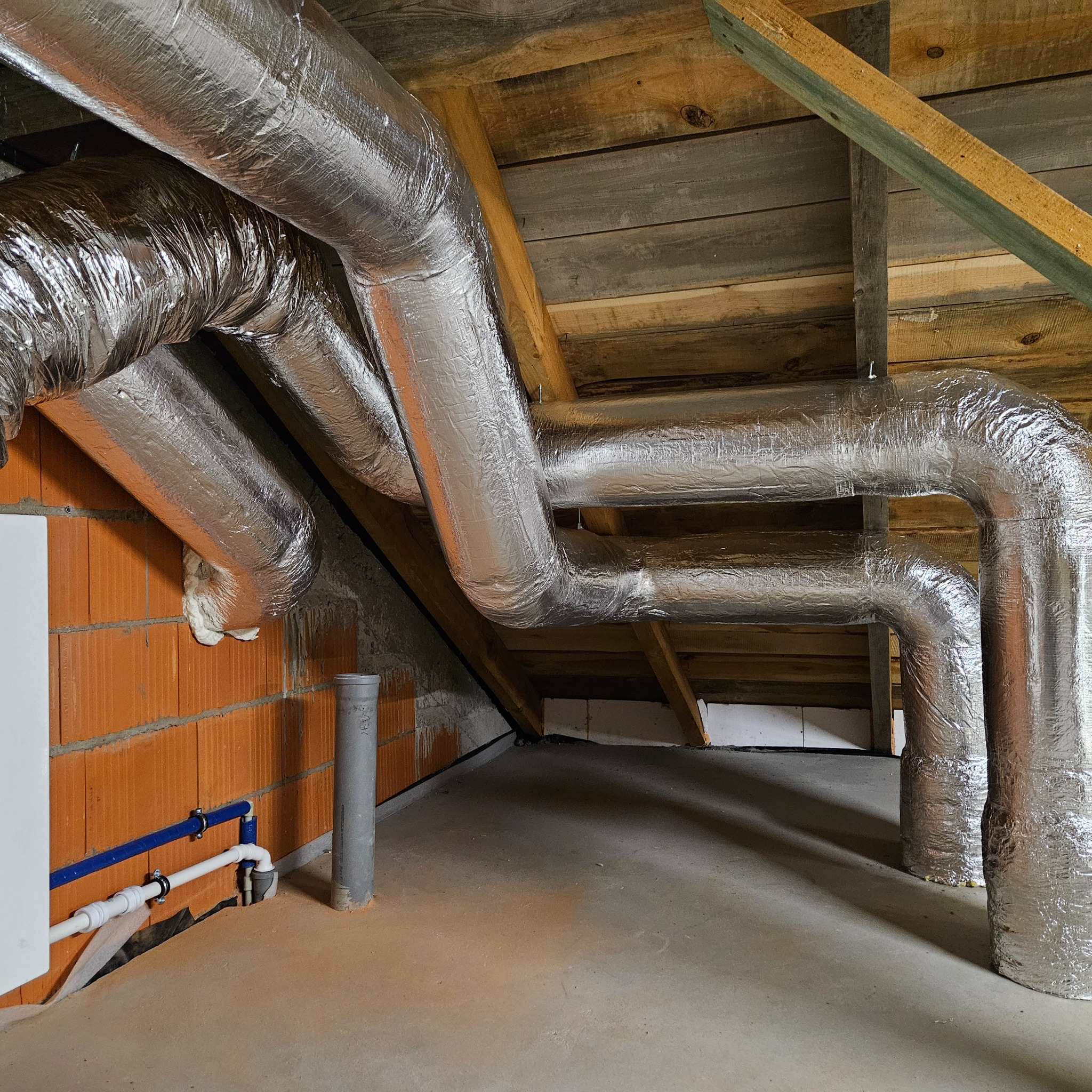 Izolowane, srebrne rury wentylacyjne w surowym pomieszczeniu poddasza, z widoczną ceglaną ścianą i drewnianą konstrukcją dachu. Instalacja w trakcie realizacji.