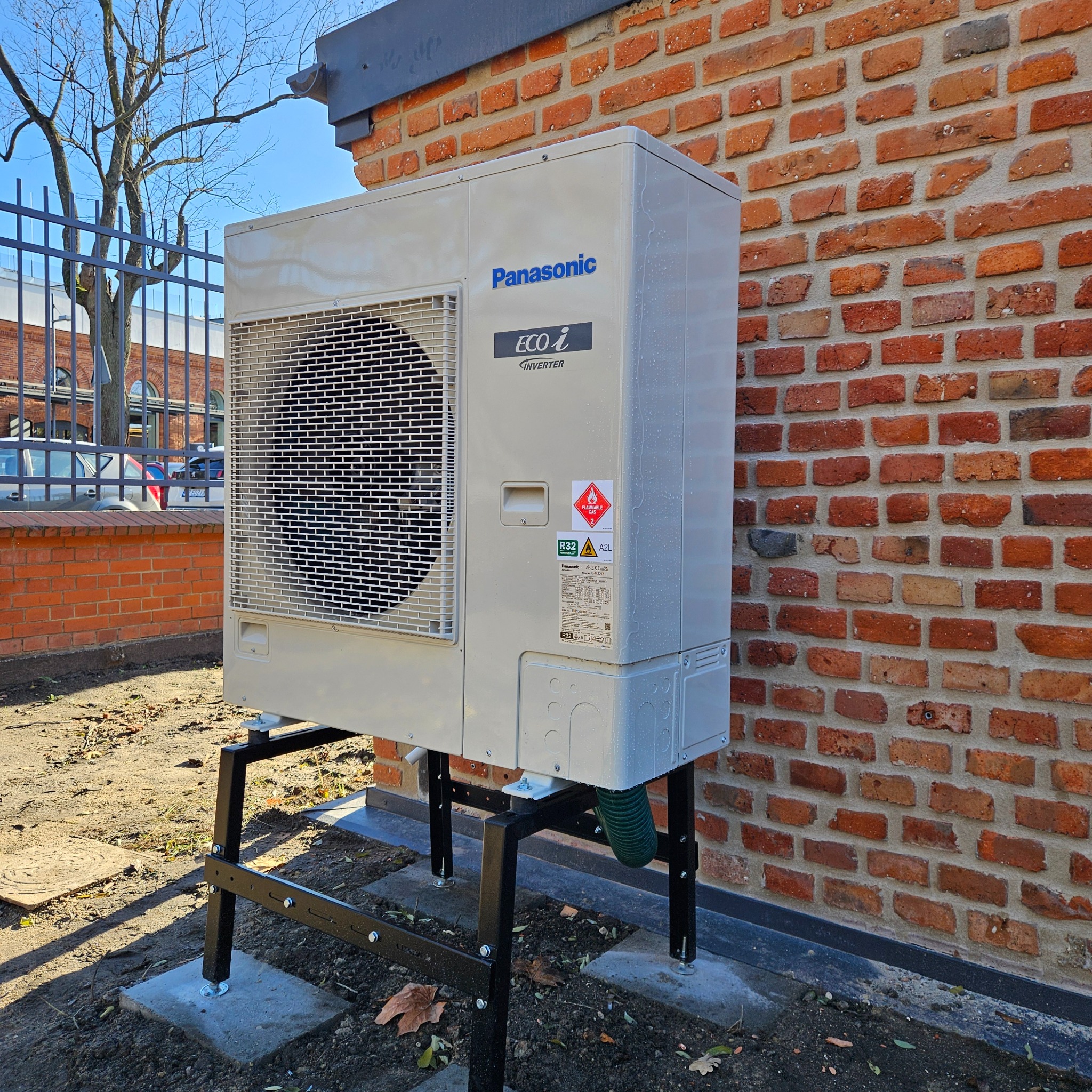 Zewnętrzna jednostka pompy ciepła Panasonic Eco i Inverter na czarnym stelażu, zamontowana przy ceglanej ścianie budynku. Widoczne elementy instalacji i otoczenie.