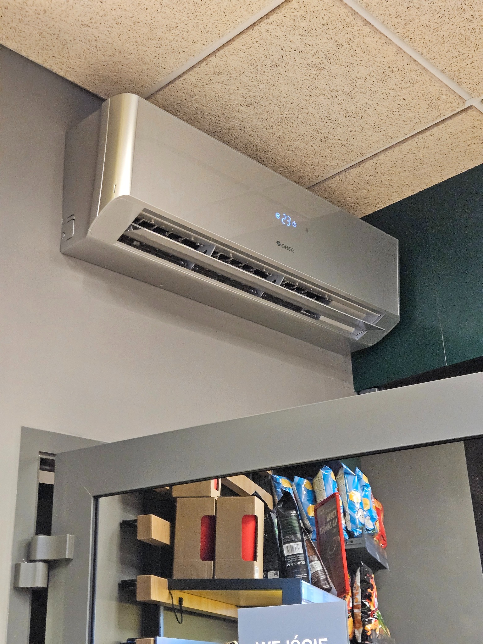 Zamontowany klimatyzator Gree na ścianie w pomieszczeniu komercyjnym, widoczny wyświetlacz temperatury 23 stopnie Celsjusza. W tle regał z produktami.