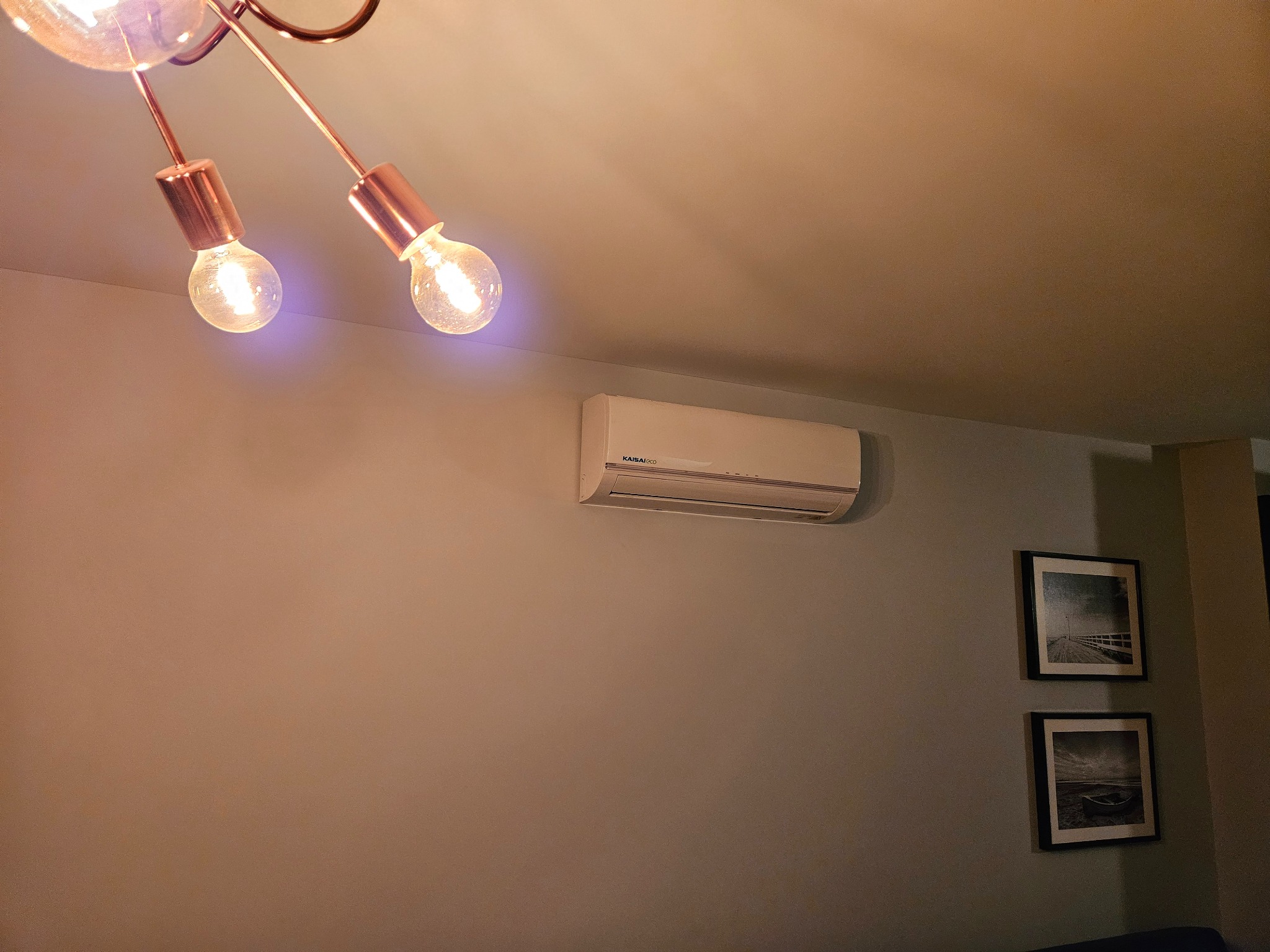 Biały klimatyzator Kaisai zamontowany na ścianie w pokoju z lampą w stylu loft i dwoma obrazami w czarnych ramkach. Widok z dołu.