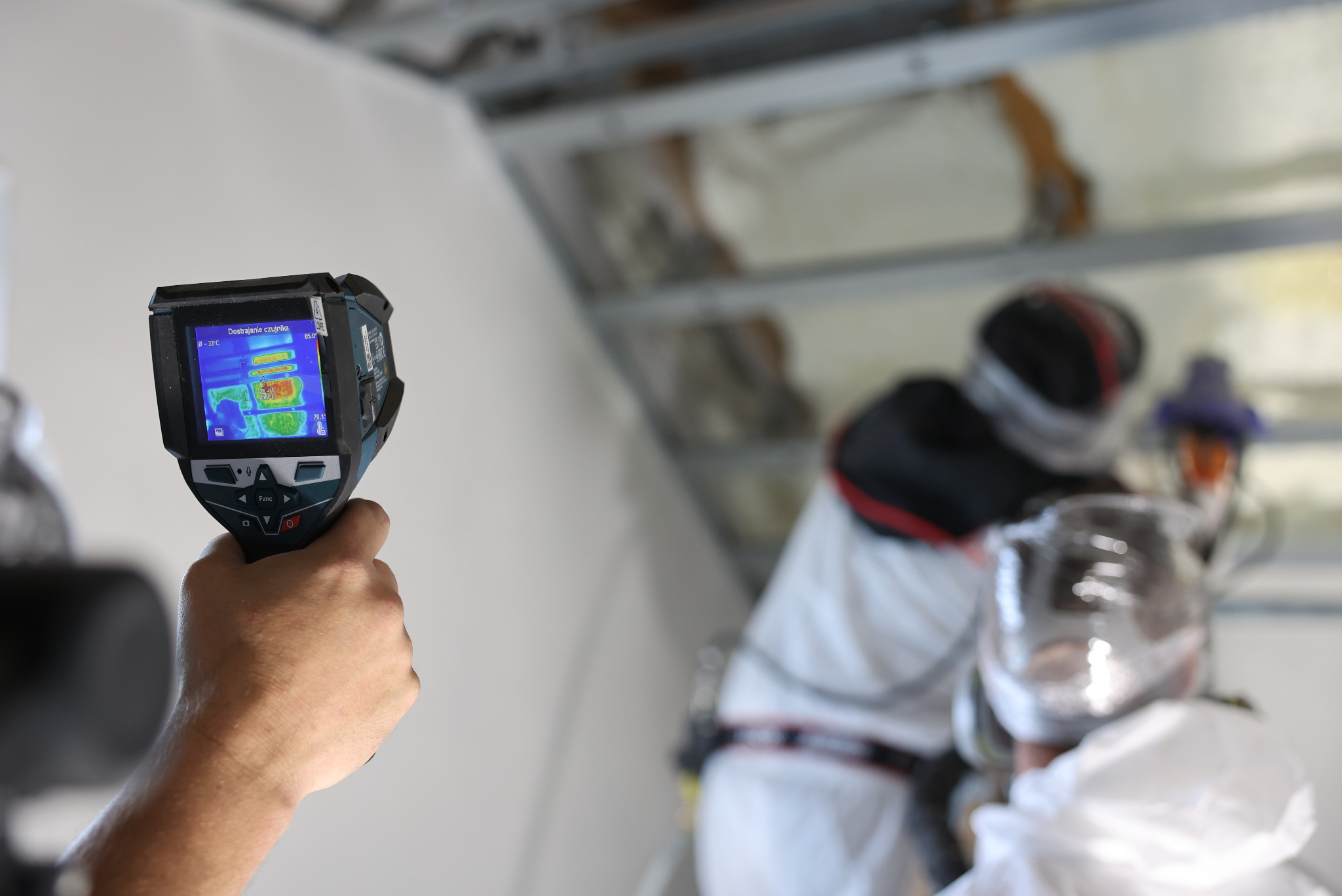 Termowizja w dłoni podczas ocieplania poddasza pianką PUR. Na ekranie termowizyjnym widoczny rozkład temperatur. Pracownik w kombinezonie ochronnym w tle.