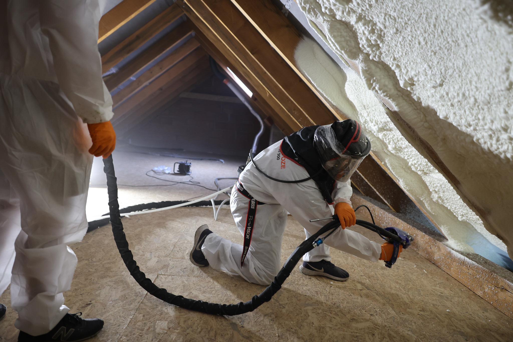Pracownik w kombinezonie natryskuje pianę izolacyjną na poddasze, widoczny fragment konstrukcji dachu i sprzęt do aplikacji.