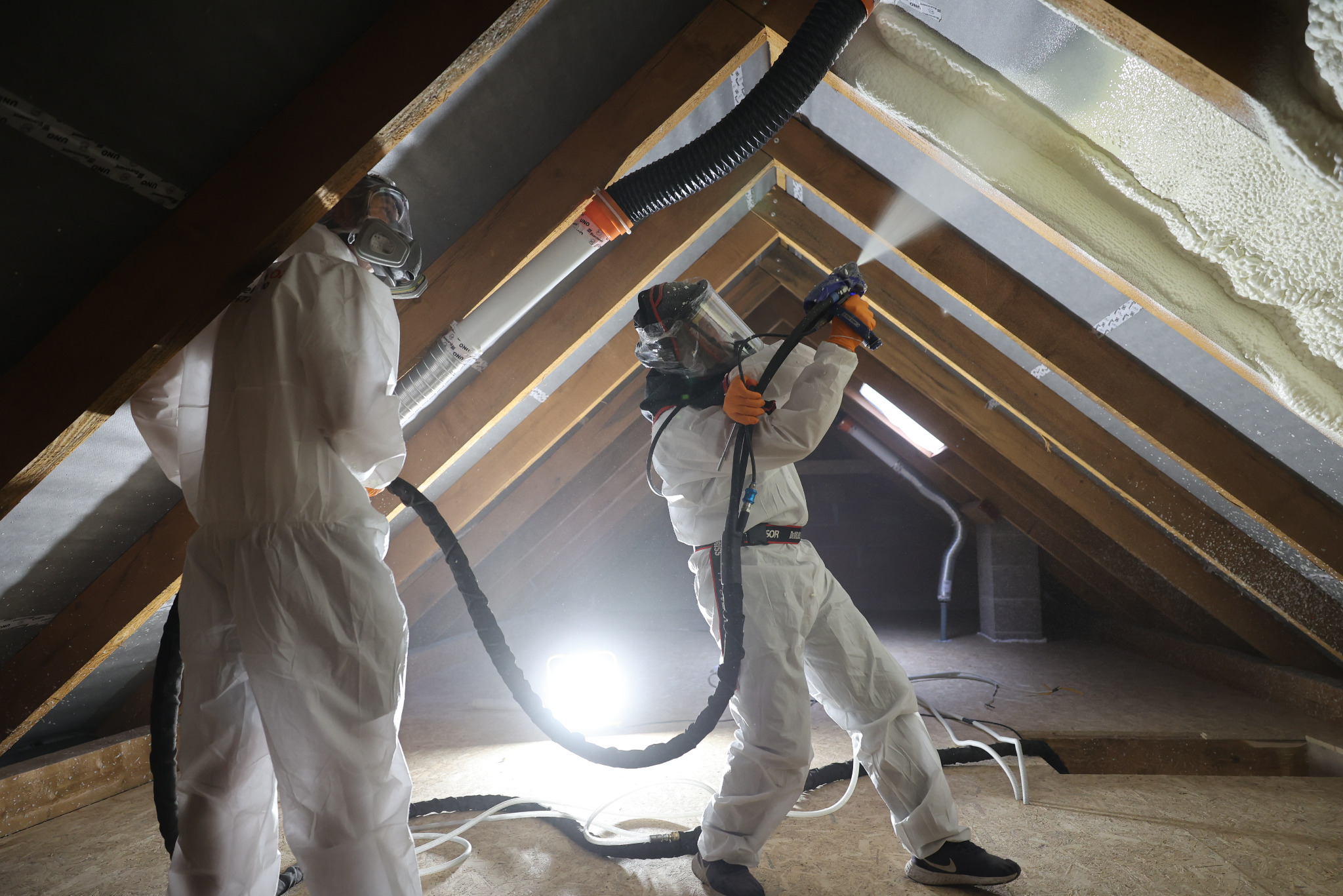 Pracownicy w kombinezonach ochronnych aplikują piankę izolacyjną na poddaszu, widoczna drewniana konstrukcja dachu i świeżo nałożona pianka.