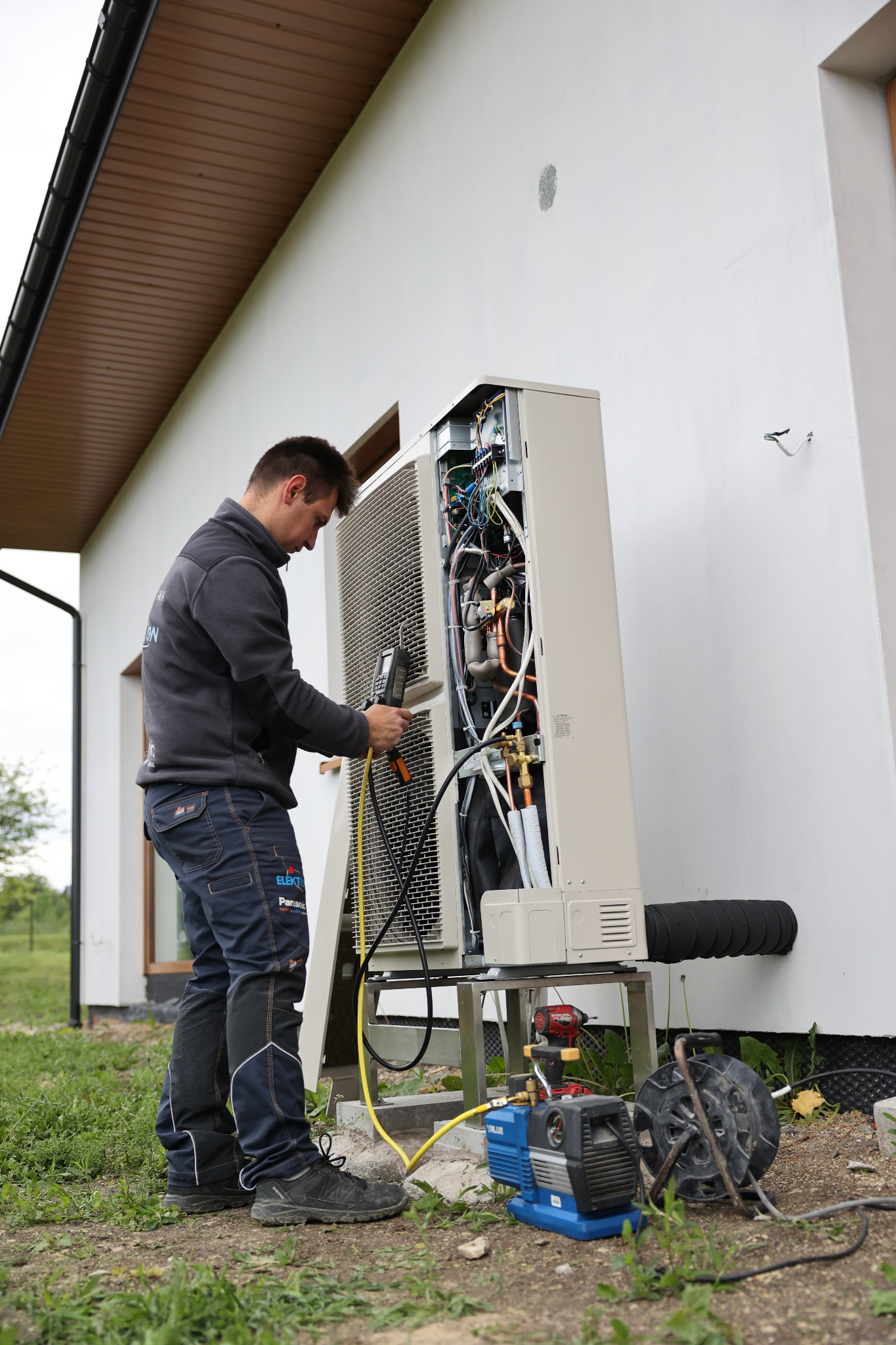 Instalator diagnozuje pompę ciepła przy budynku. Widoczne narzędzia i otwarte urządzenie, ukazujące wewnętrzne komponenty. Praca w toku.