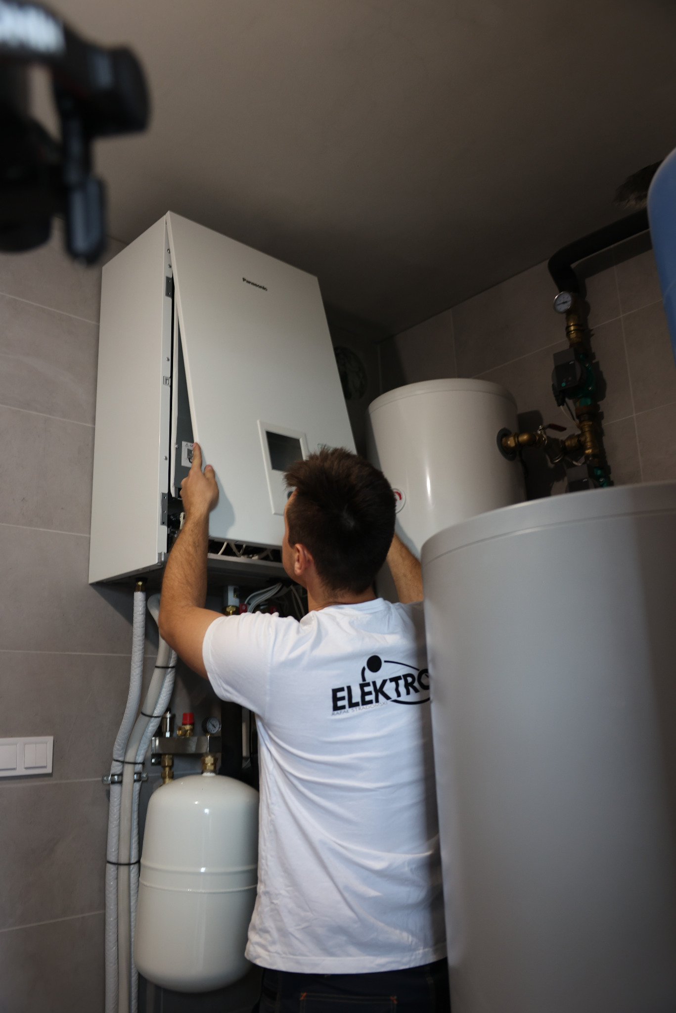 Instalator w białej koszulce serwisuje biały piec gazowy Panasonic w kotłowni z szarymi płytkami, widoczne rury i zbiorniki.