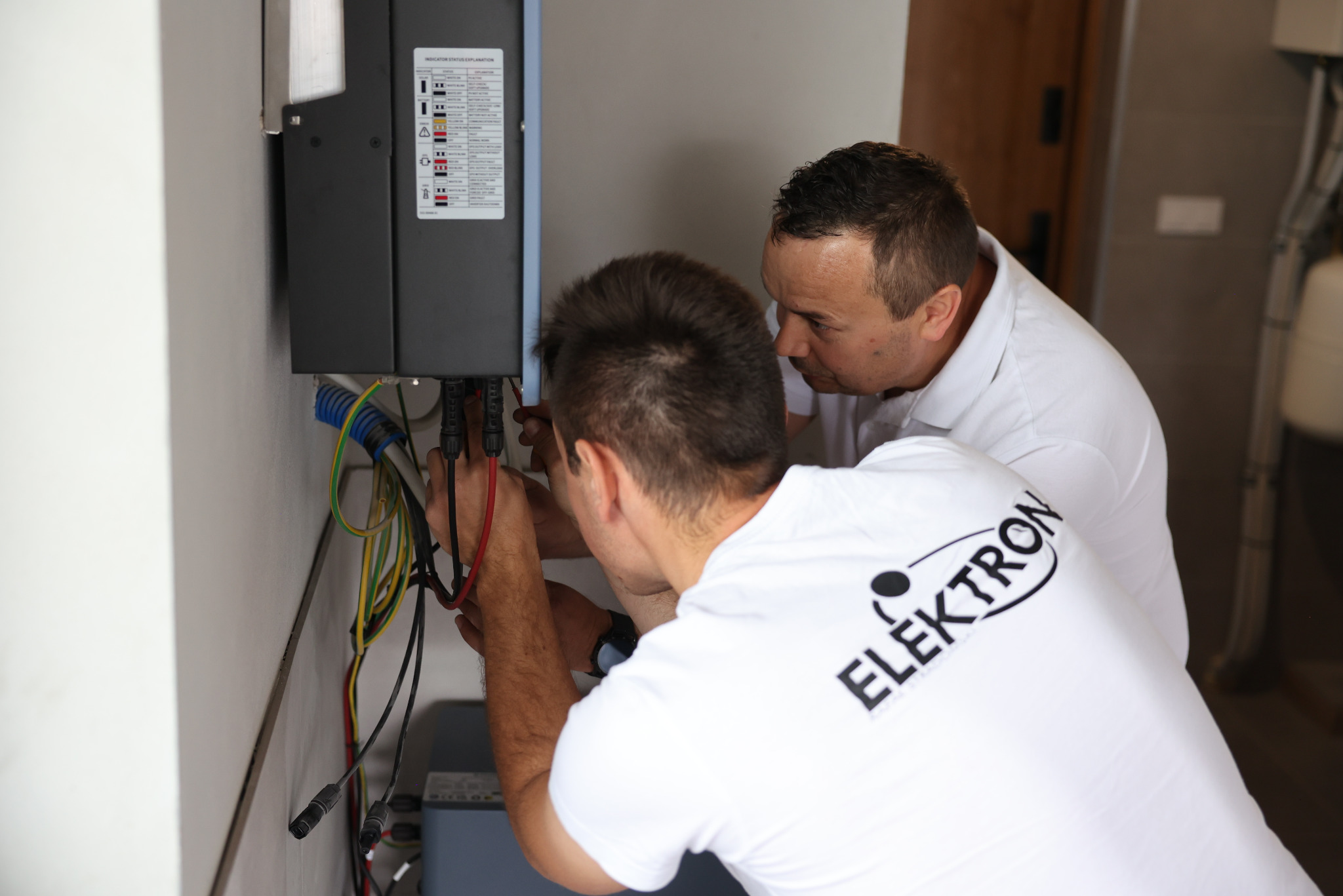 Dwóch instalatorów w białych koszulkach z logo firmy podłącza kable do urządzenia zamontowanego na ścianie. Widoczne przewody w kolorach: czerwonym, żółtym i niebieskim.