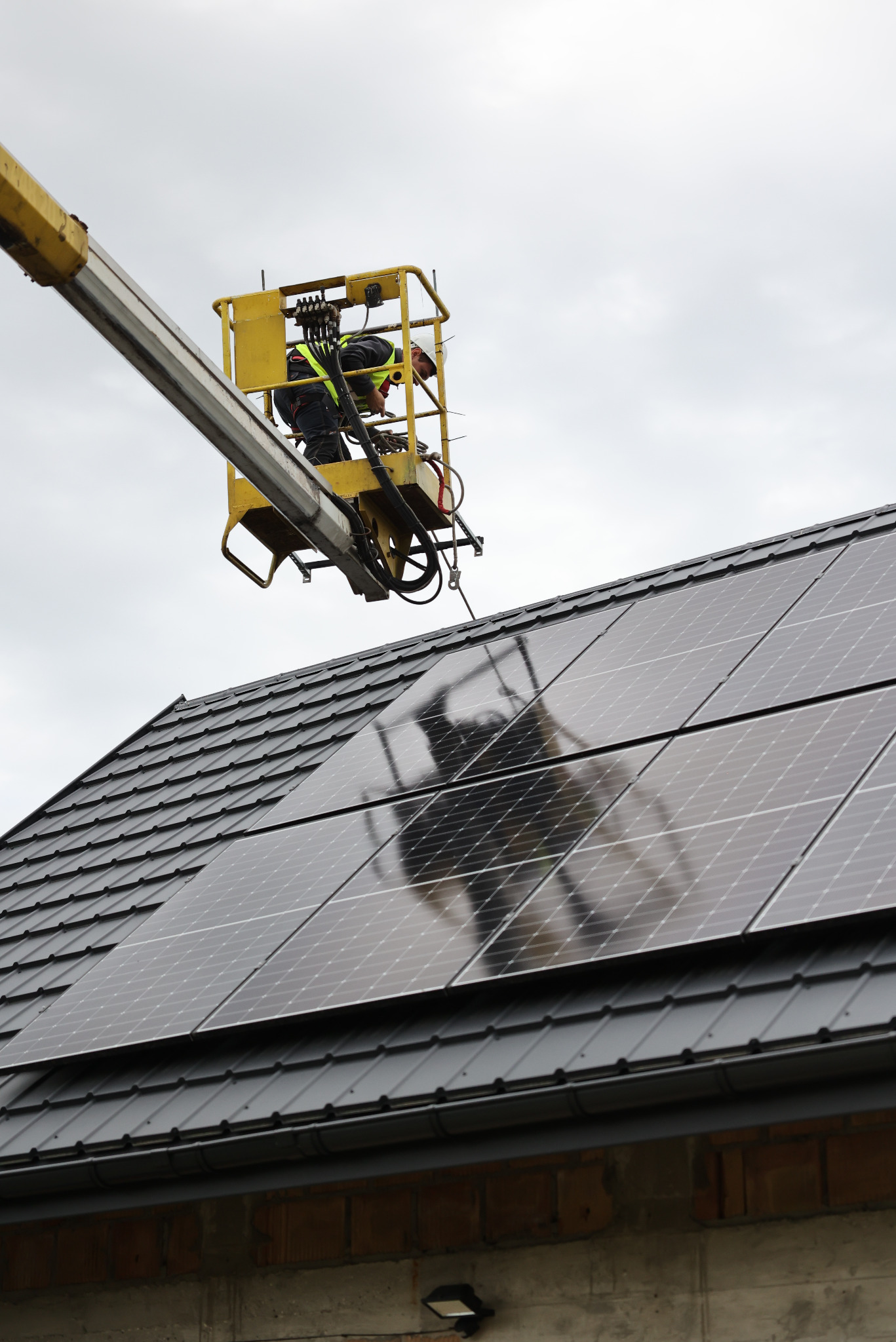 Montaż paneli słonecznych na dachu z użyciem podnośnika. Pracownik w kasku i kamizelce. Widoczne panele, dach pokryty dachówką, fragment budynku.